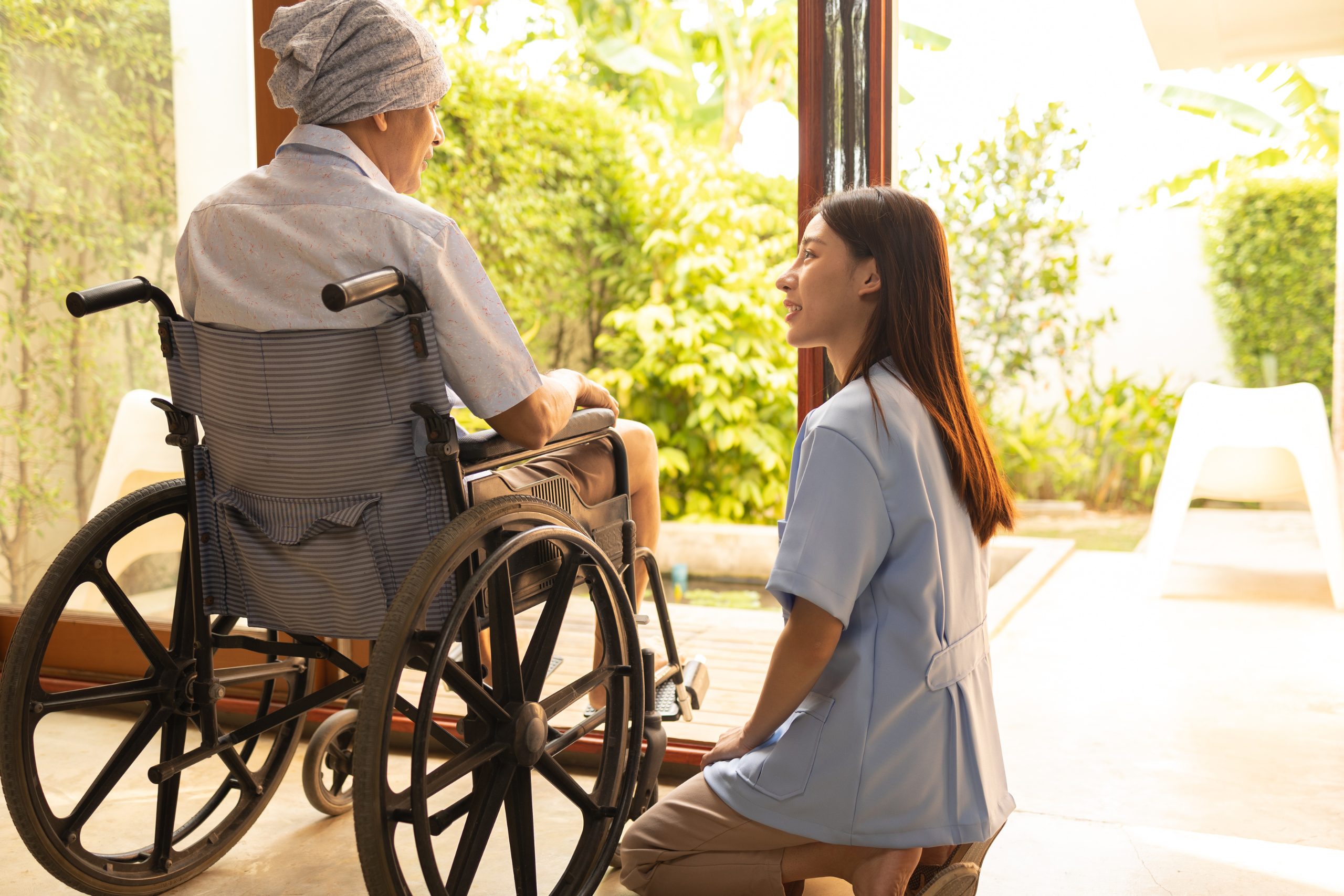 A caring nurse in blue scrubs kneels beside a senior patient in a wheelchair, engaging in a warm, attentive conversation, illustrating the compassionate advocacy and communication in nursing home care.
