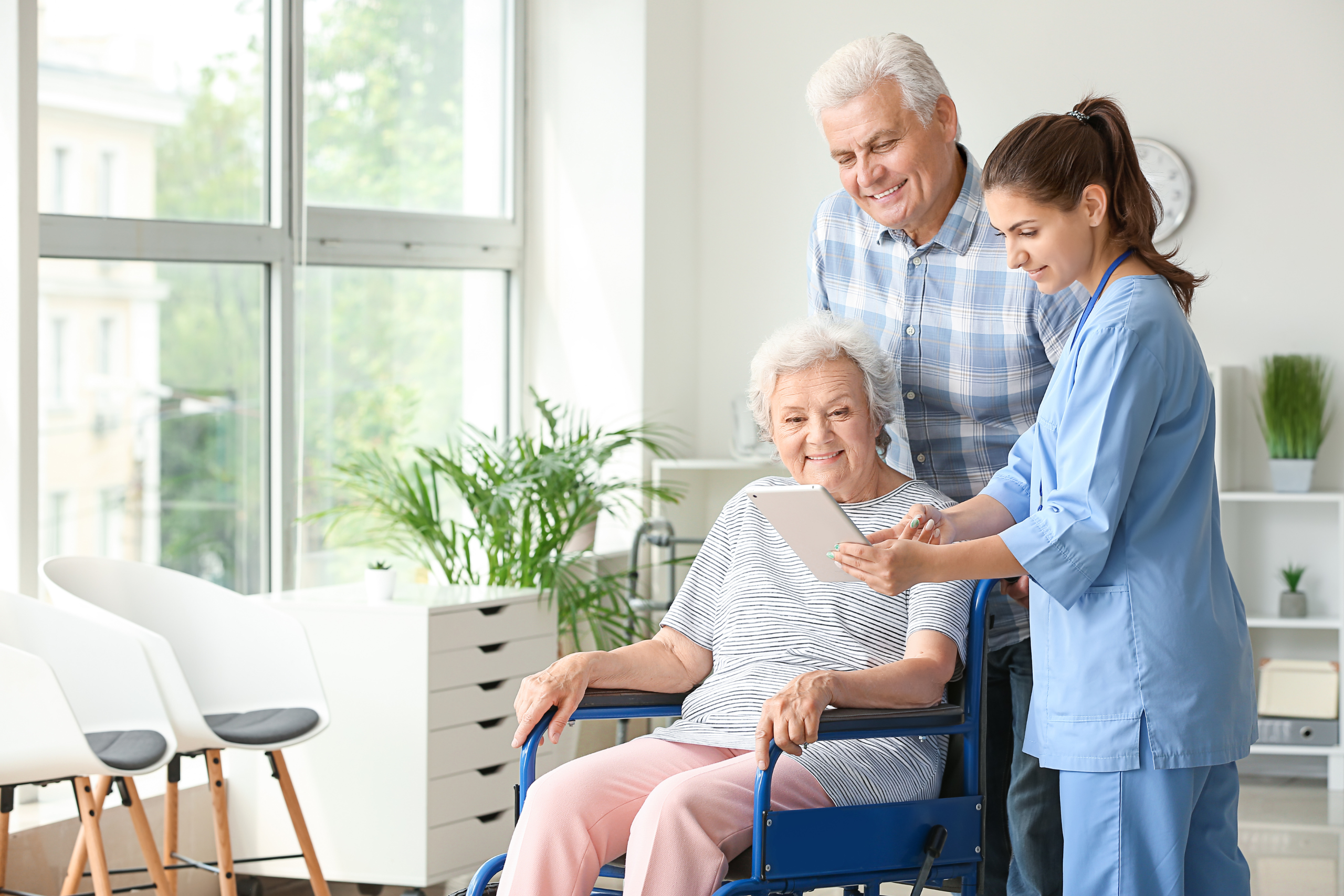 Elderly woman in a wheelchair smiling while looking at a tablet with a healthcare worker, as a senior man stands behind her, depicting a family involved in care decisions, emphasizing the theme of being an advocate for loved ones.