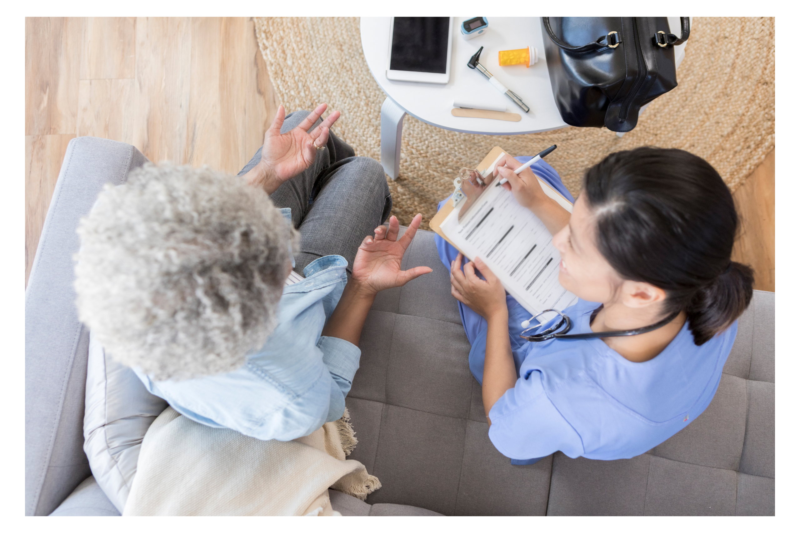 Overhead view of a healthcare professional in blue attire taking notes while attentively listening to an elderly woman gesturing during a conversation, symbolizing effective communication in nursing home advocacy.