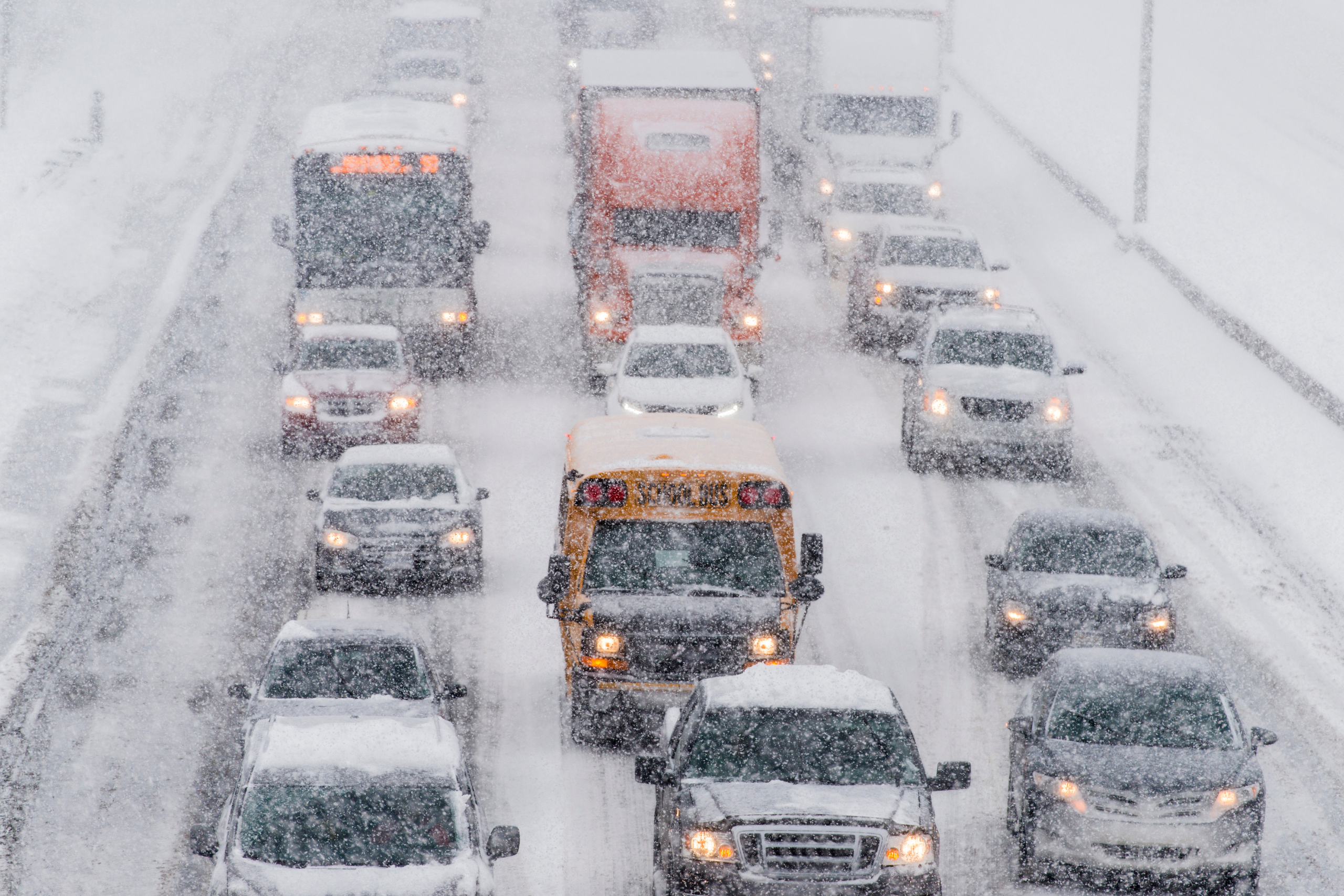 Aerial view of a busy highway with a diverse array of vehicles, including cars, trucks, and a school bus, cautiously navigating through a heavy snowfall, emphasizing the need for careful driving in winter weather conditions.