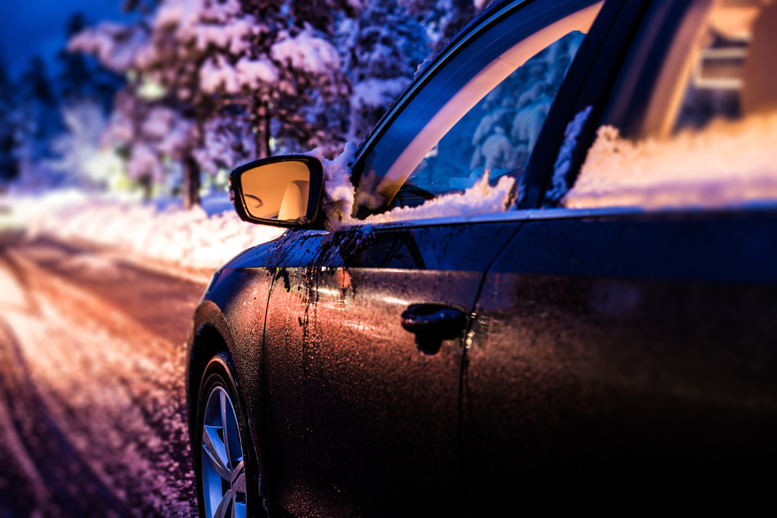 Black car parked on a snowy road at dusk, with the warm glow of street lights reflecting off its shiny surface, highlighting the importance of vehicle safety during winter conditions.