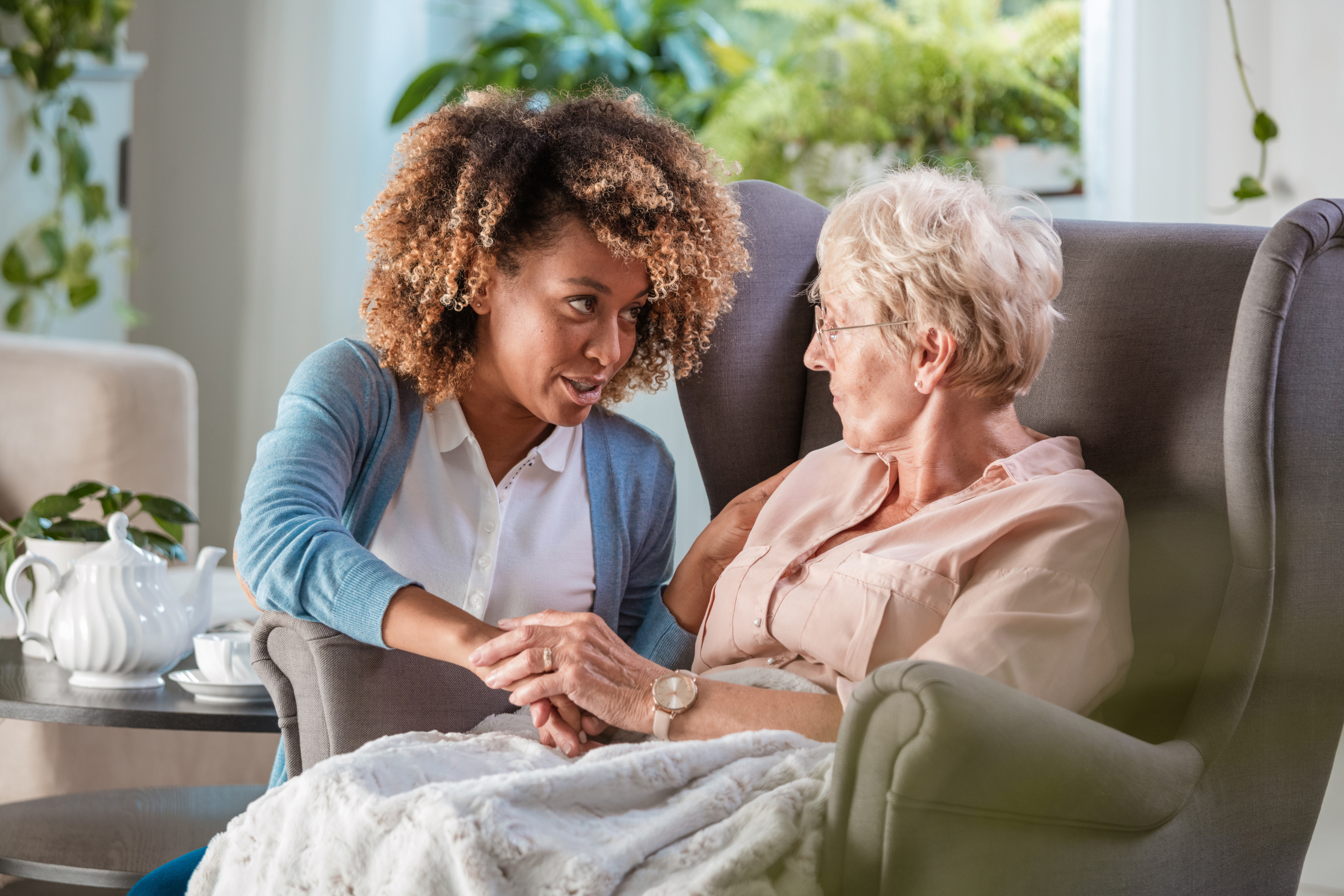 A compassionate young woman holds hands with an elderly lady while engaging in a heartfelt conversation, showcasing a moment of understanding and personal care, aligning with the concept of being an expert on your loved one's needs and well-being.