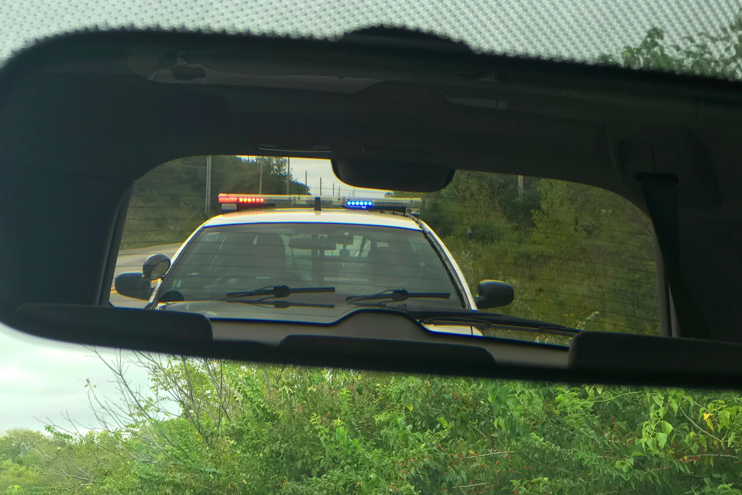 View from the inside of a car through the rearview mirror, showing a police car with flashing lights behind, suggesting the driver may have been pulled over.