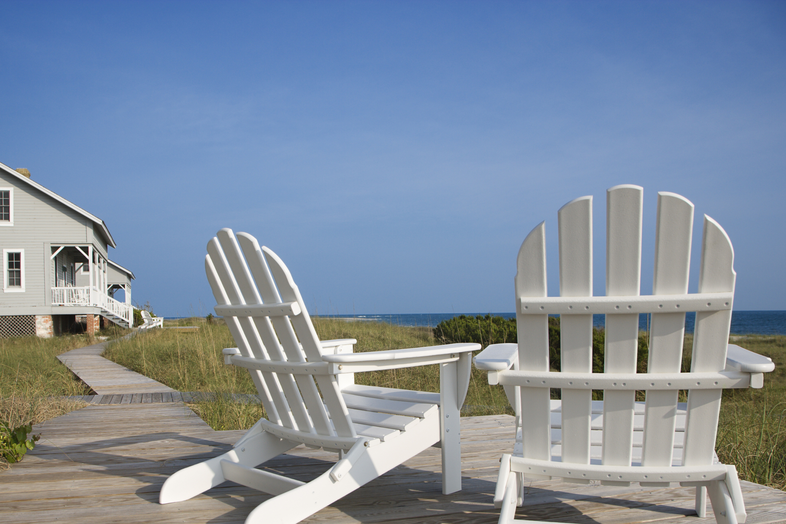 Two white Adirondack chairs on a wooden walkway with a house and ocean in the background.