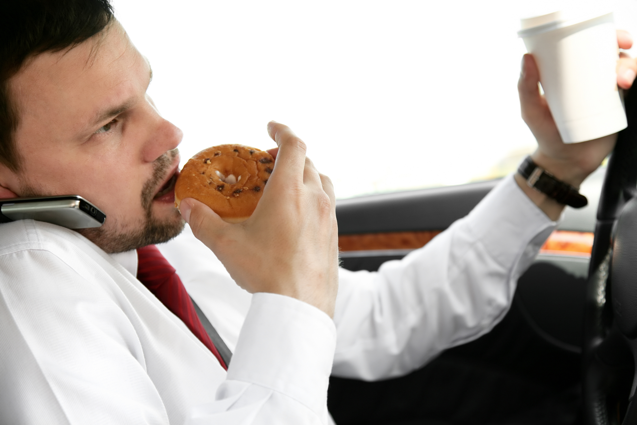A man in a white shirt and red tie is seen multitasking while driving. He is biting into a donut with his right hand, holding a coffee cup in his left hand, and talking on a cell phone wedged between his shoulder and ear.