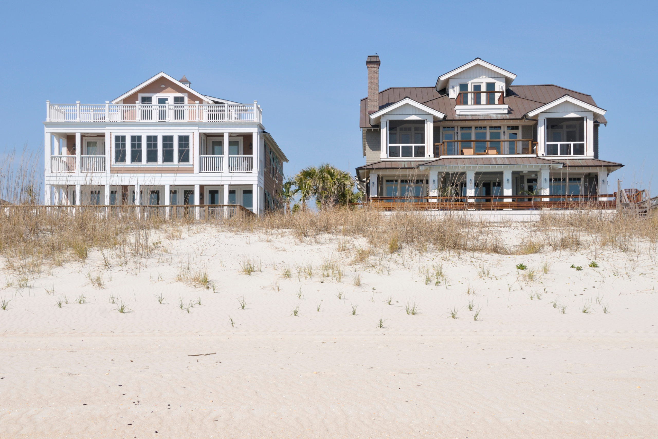 Two beachfront houses on a sandy dune with clear blue skies.