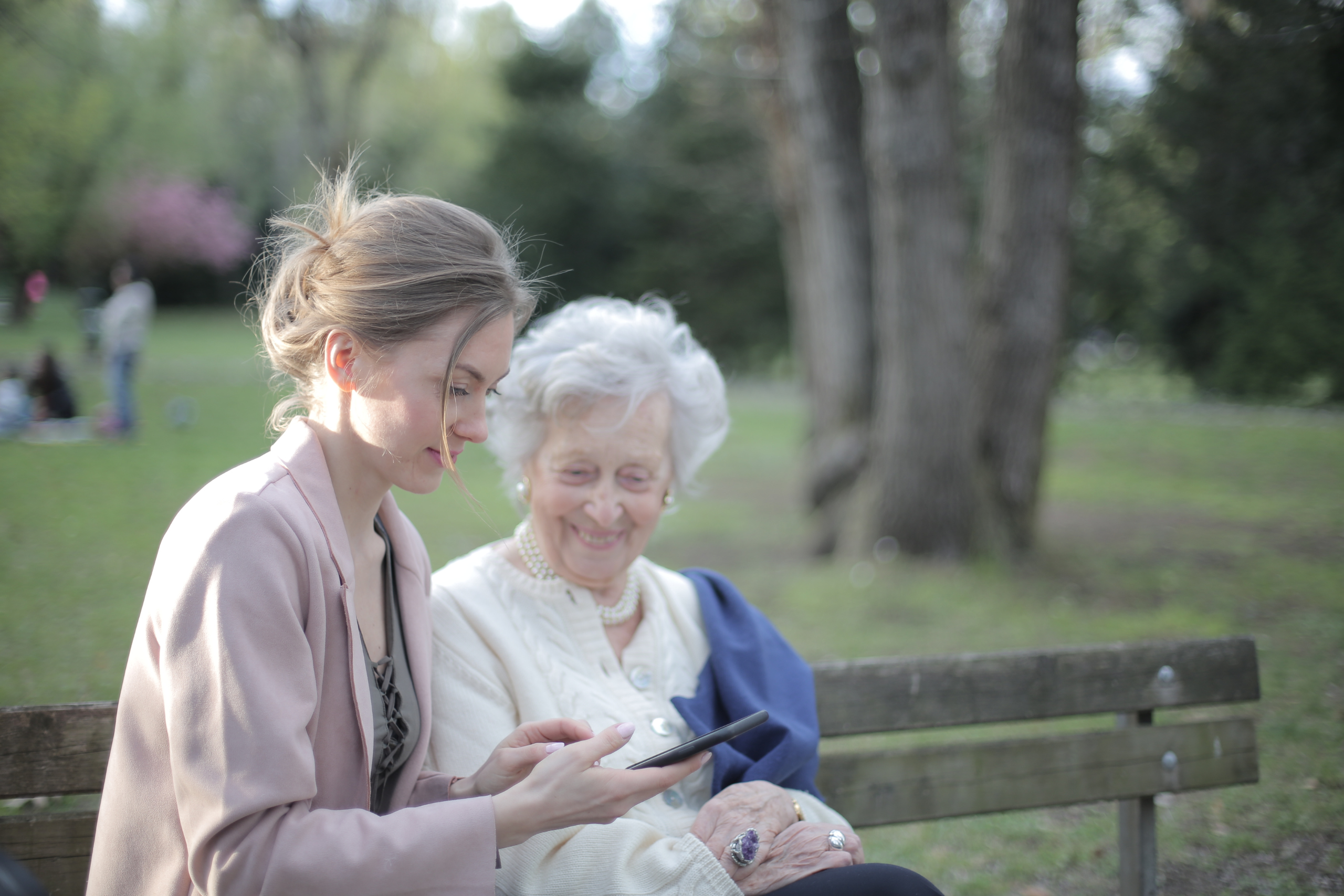A younger woman and an older woman are seated on a bench in a park, sharing a moment of joy as the younger one shows something on a smartphone to the older woman. The older woman, who is smiling, appears to be enjoying the interaction or content being shown. The background is softly blurred, indicating a peaceful outdoor setting with trees and possibly other park-goers in the distance.