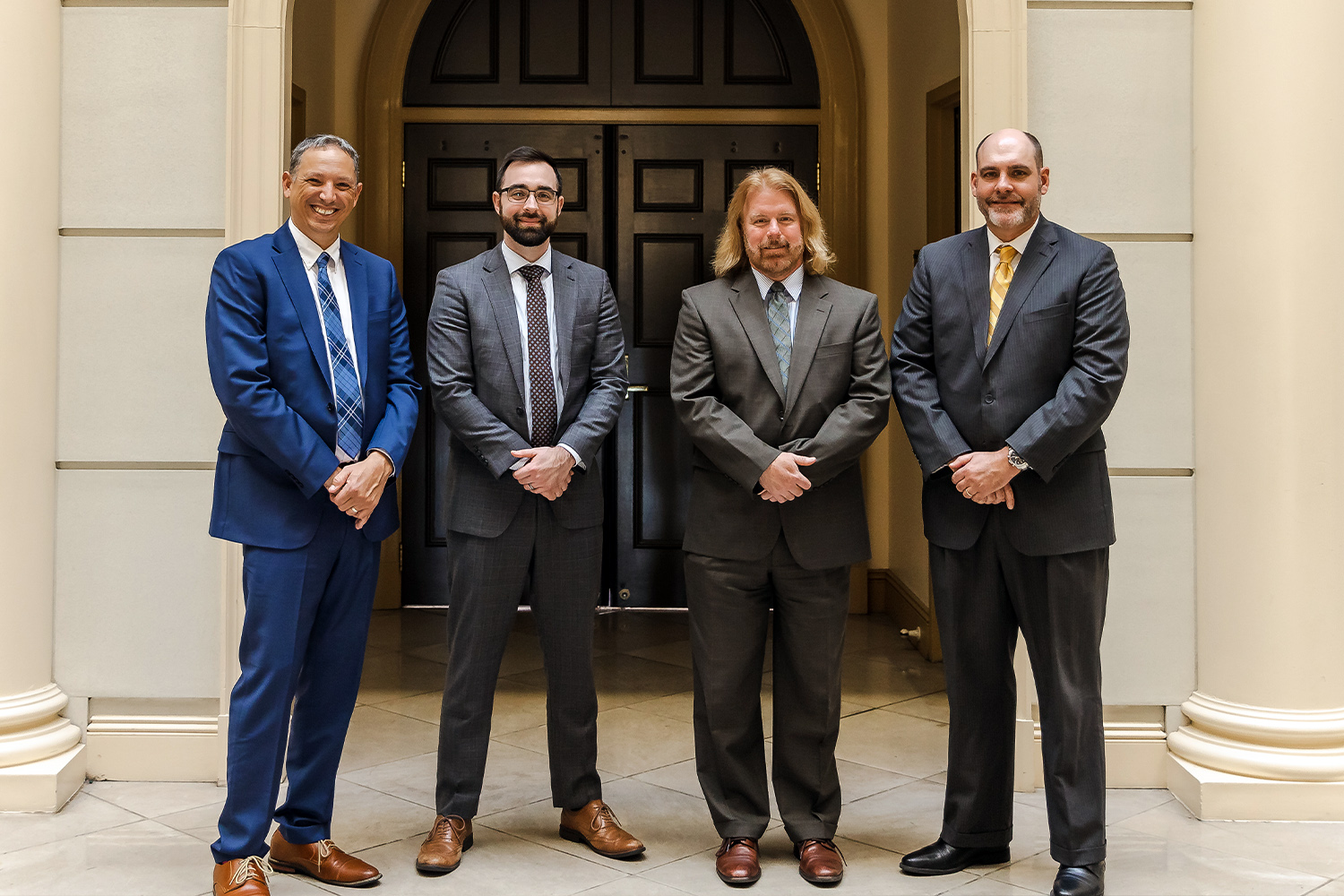 Four businessmen in suits stand together in a hallway, smiling confidently.