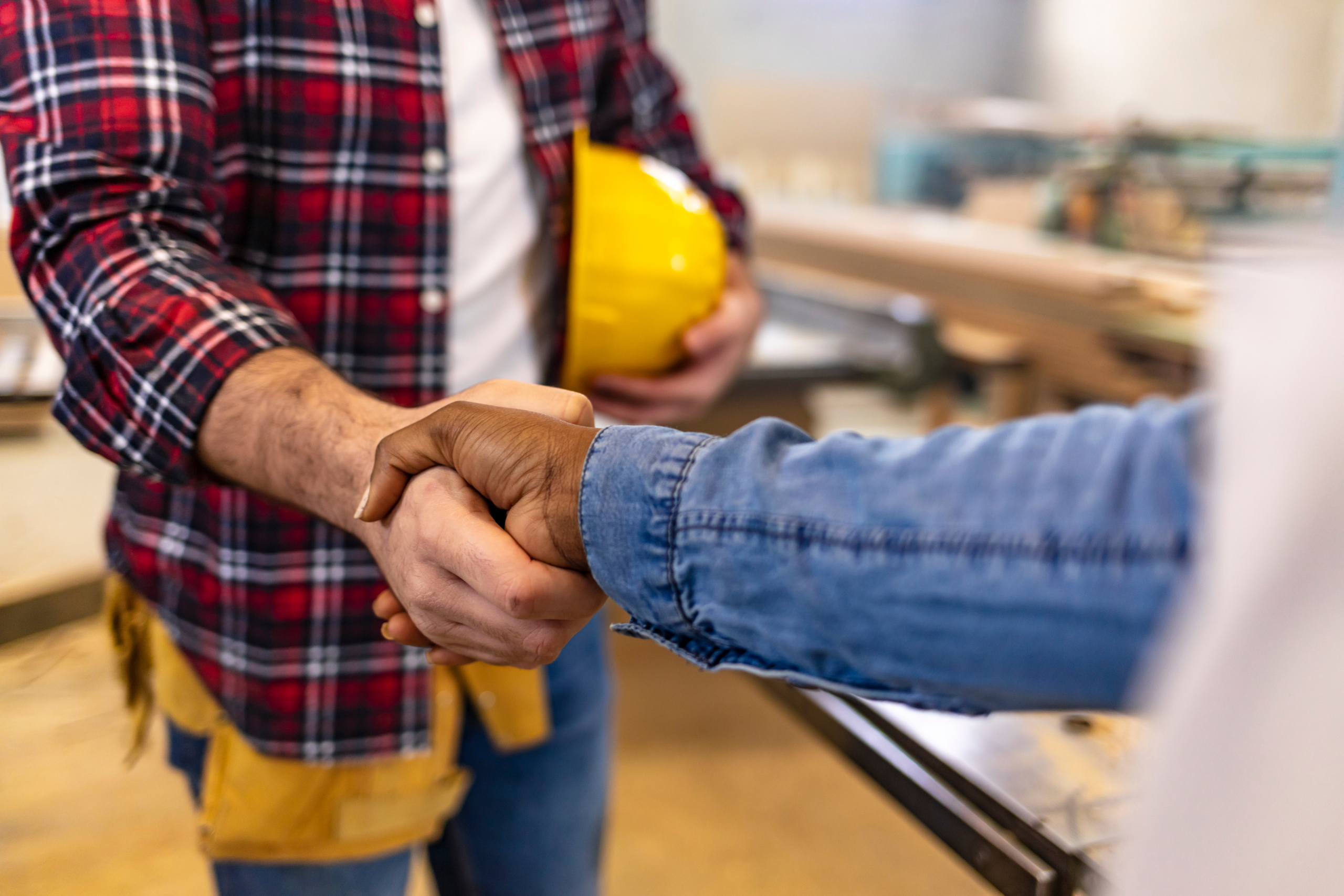 Close-up of two people shaking hands in a workshop, one holding a yellow hard hat.