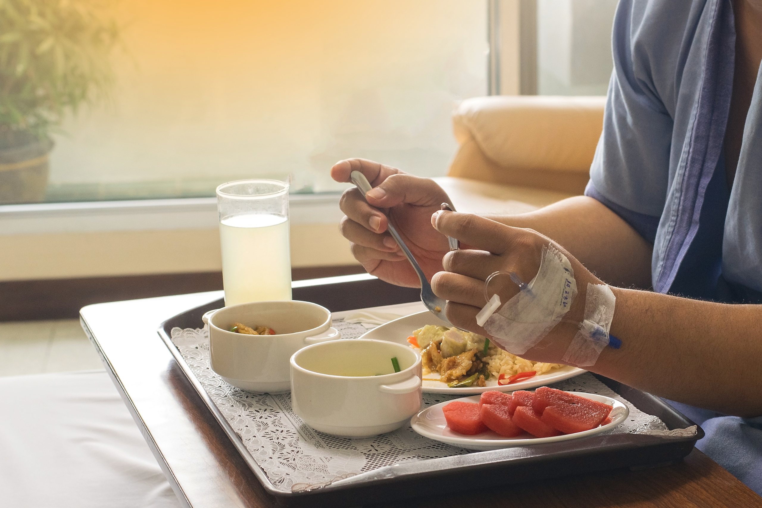 Person in hospital gown eating a nutritious meal on a tray, with water and fresh fruit, emphasizing proper care in nursing homes.