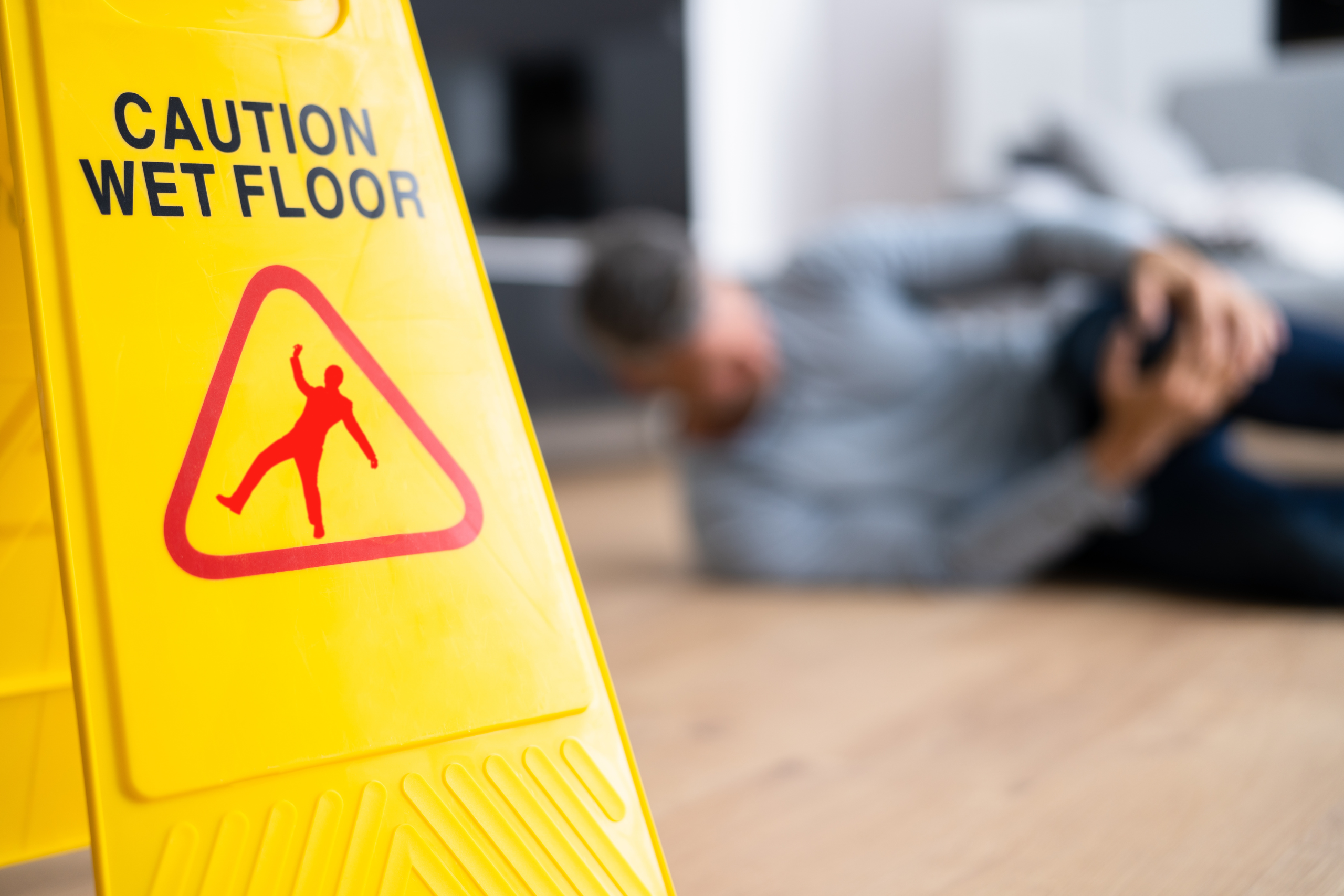 Foreground focus on a 'Caution Wet Floor' sign with a blurry background of a man holding his knee on the floor.