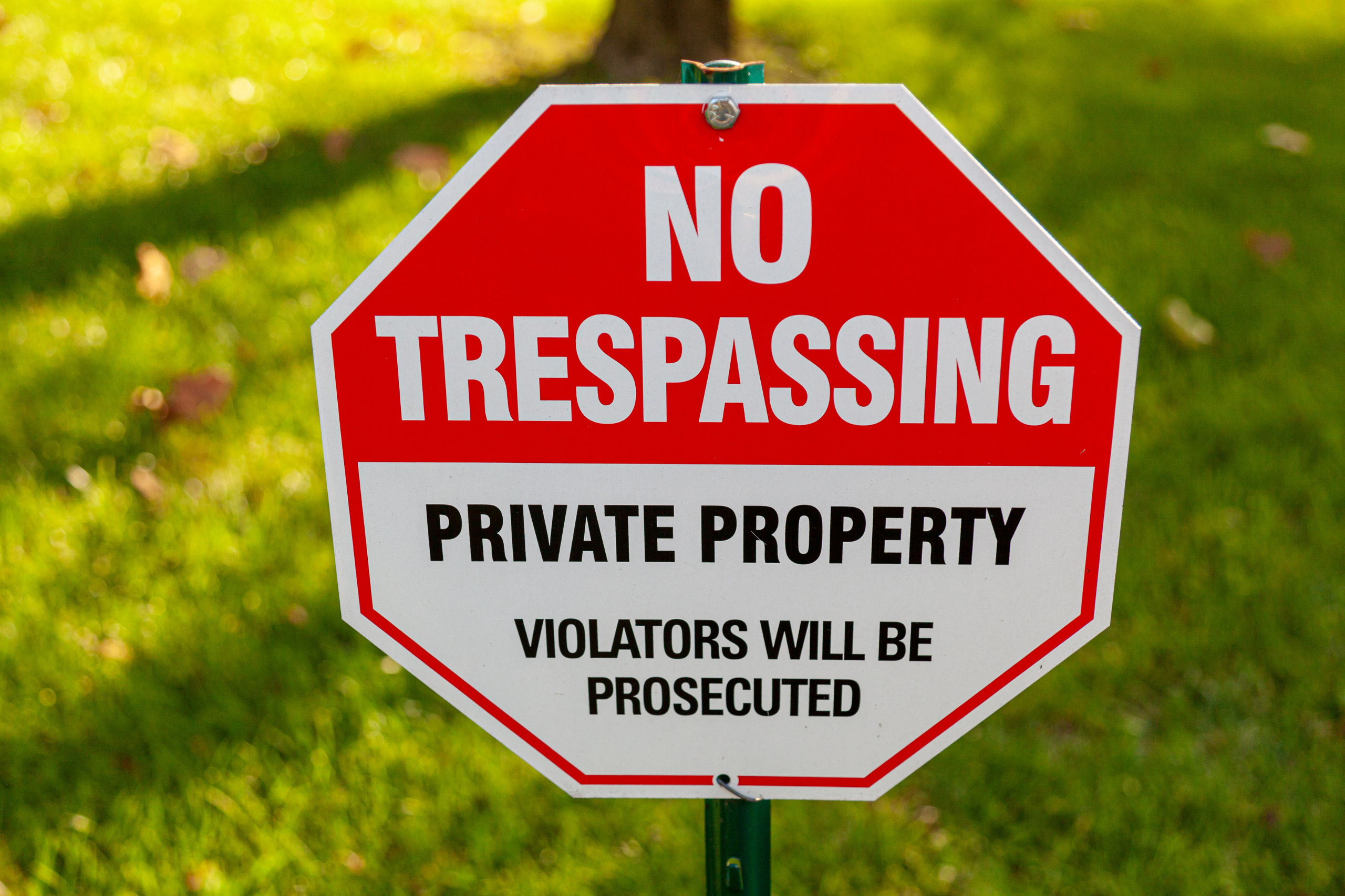 Red and white 'No Trespassing' sign on a green background stating 'Private Property, Violators will be prosecuted.'
