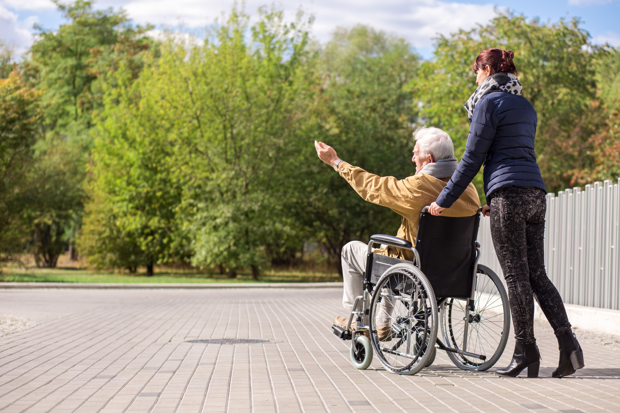 Elderly man in wheelchair outside with woman pointing at park.