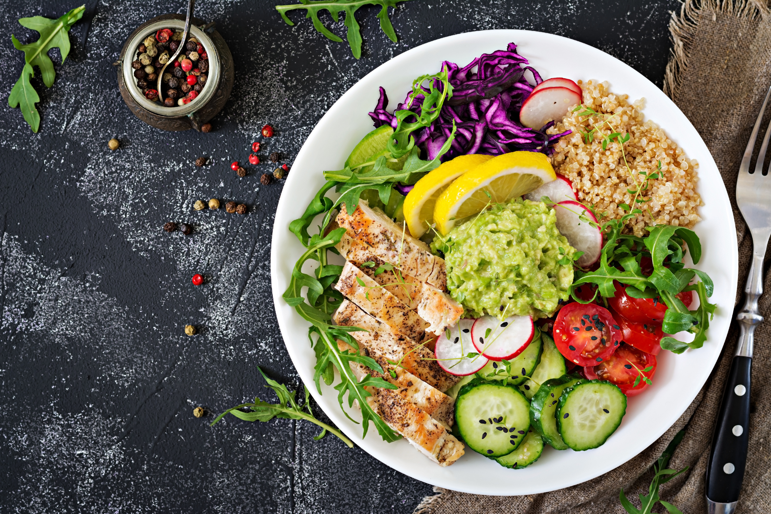 Colorful balanced meal plate with grilled chicken, quinoa, guacamole, and fresh vegetables, served on a dark background.