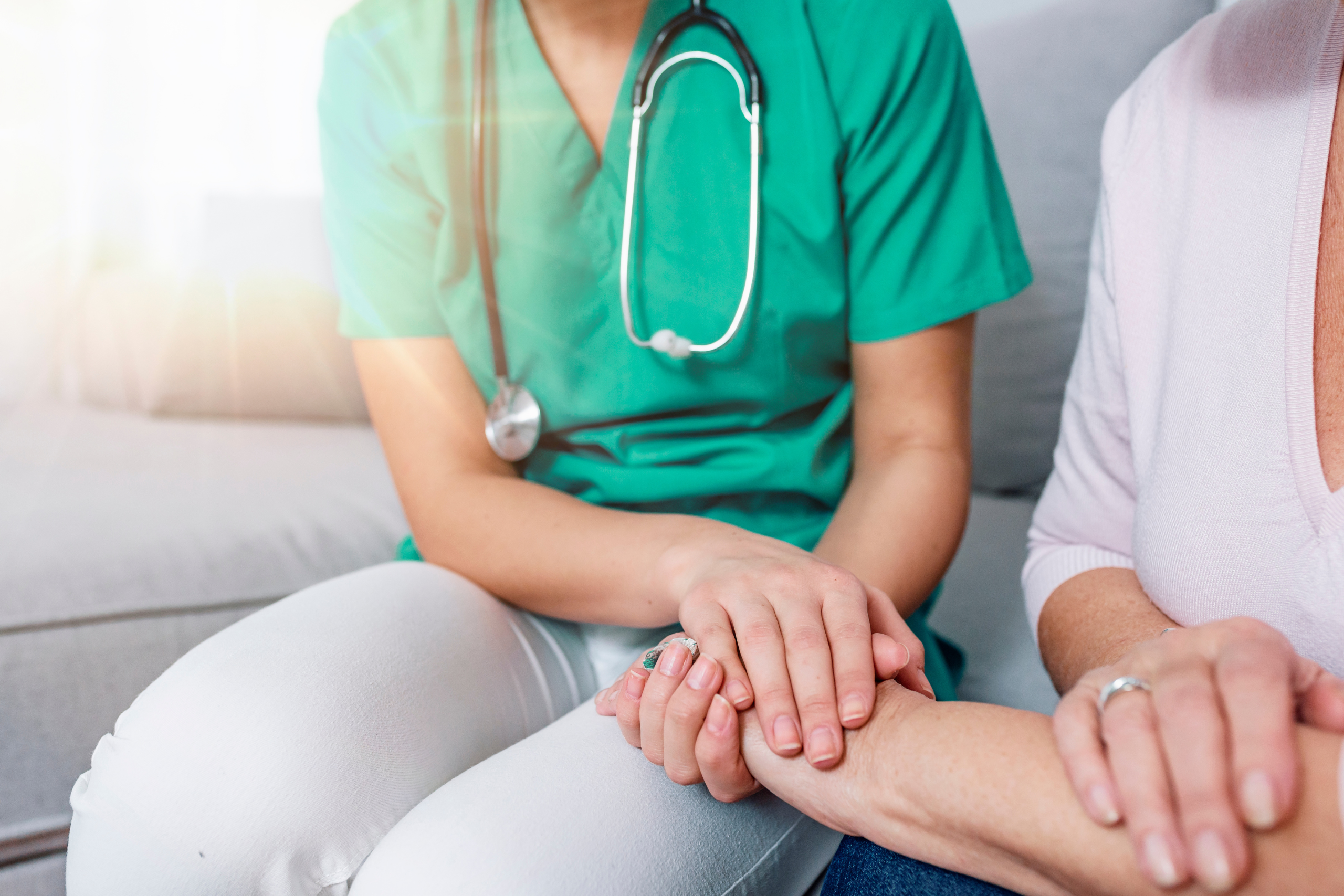 A healthcare worker in green scrubs holds a patient's hand, offering support in a comfortable setting.