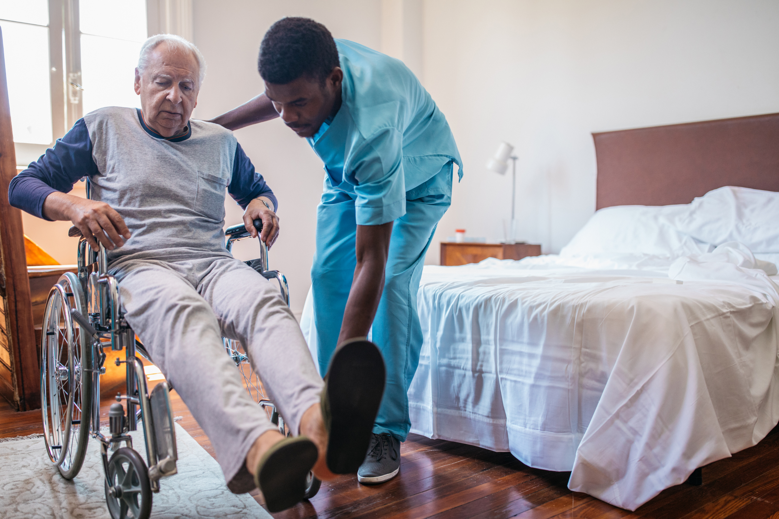 A caregiver assists an elderly man in a wheelchair near a bed in a brightly lit room.