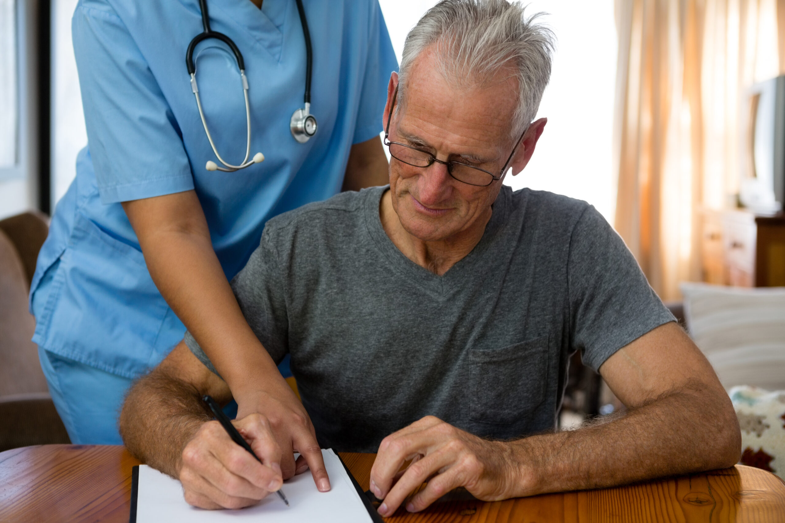 An elderly man with glasses signs a document while a nurse in blue scrubs assists by pointing at the clipboard.