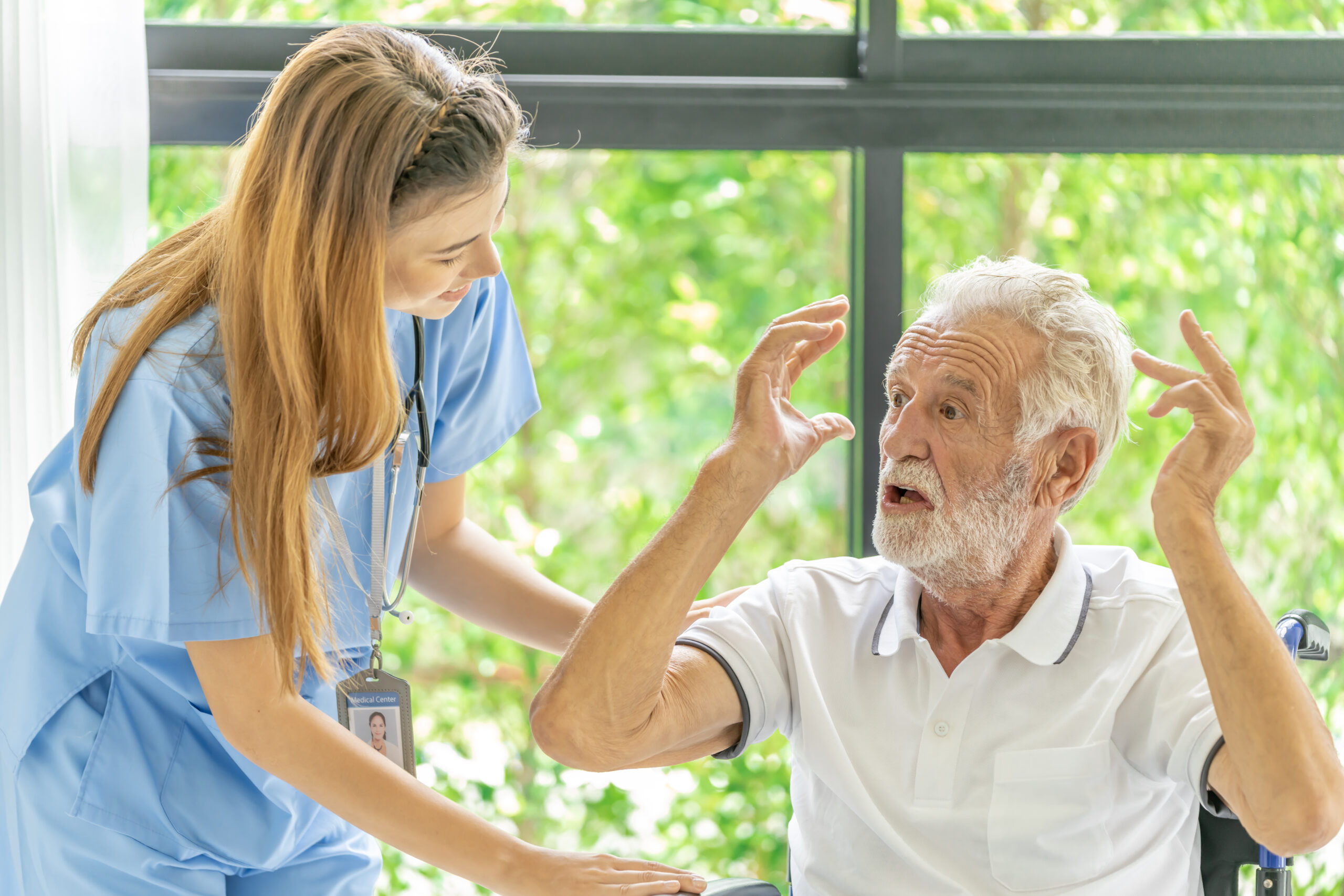 A concerned elderly man in a wheelchair gestures while speaking with a nurse in blue scrubs, illustrating communication in a healthcare setting.