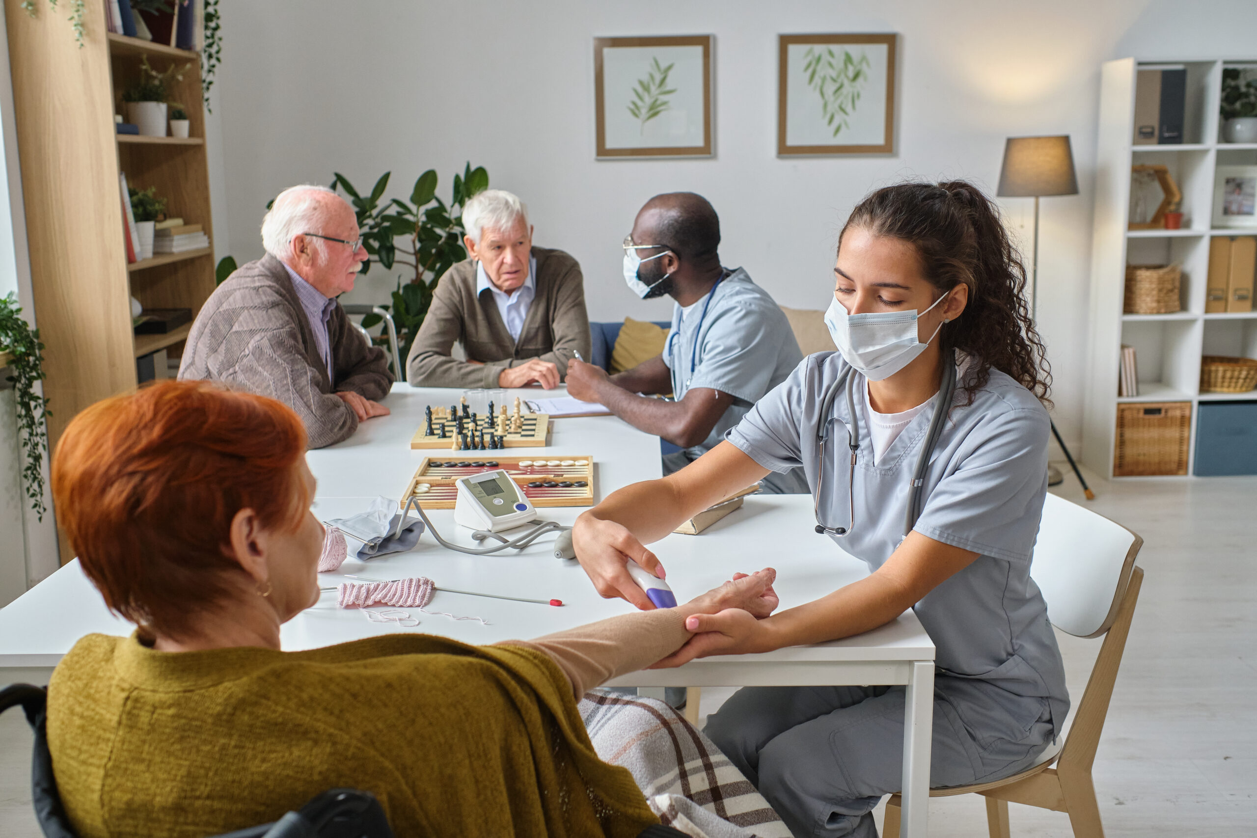 A nurse in scrubs checks a senior woman's blood pressure, while other elderly residents and staff interact at a nearby table in a nursing home.