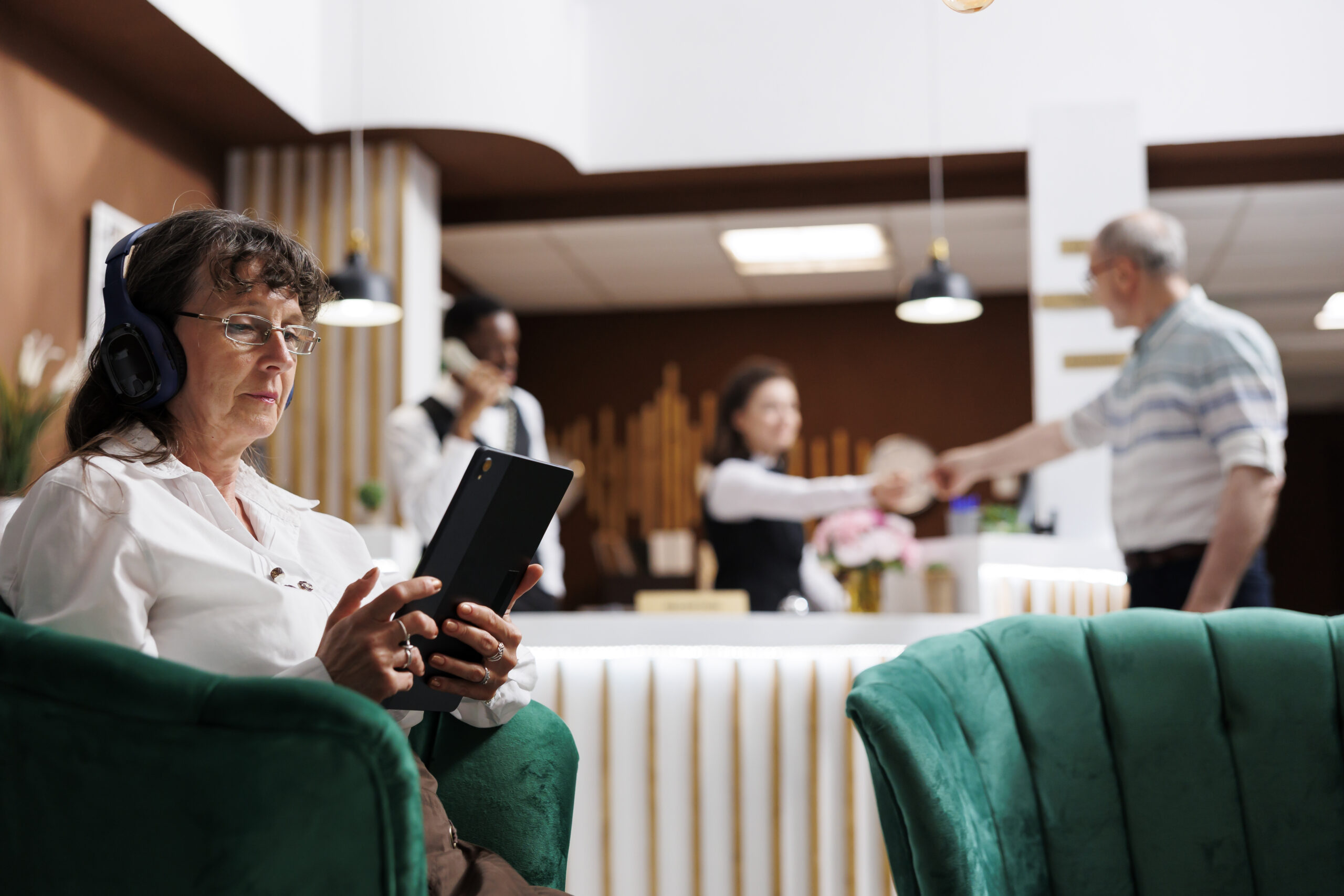 Woman wearing headphones and using a tablet in a lobby, with staff interacting in the background, emphasizing staying informed about key matters.