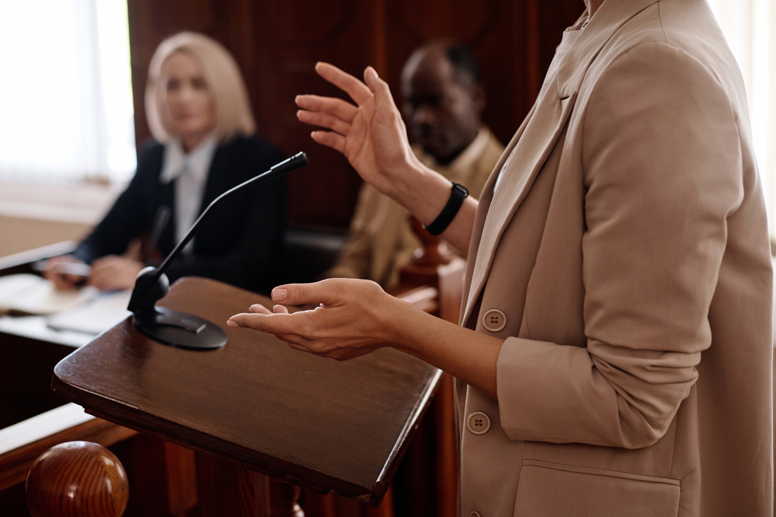 Close-up of a woman gesturing while speaking at a courtroom podium, with two blurred figures, possibly legal professionals, in the background.