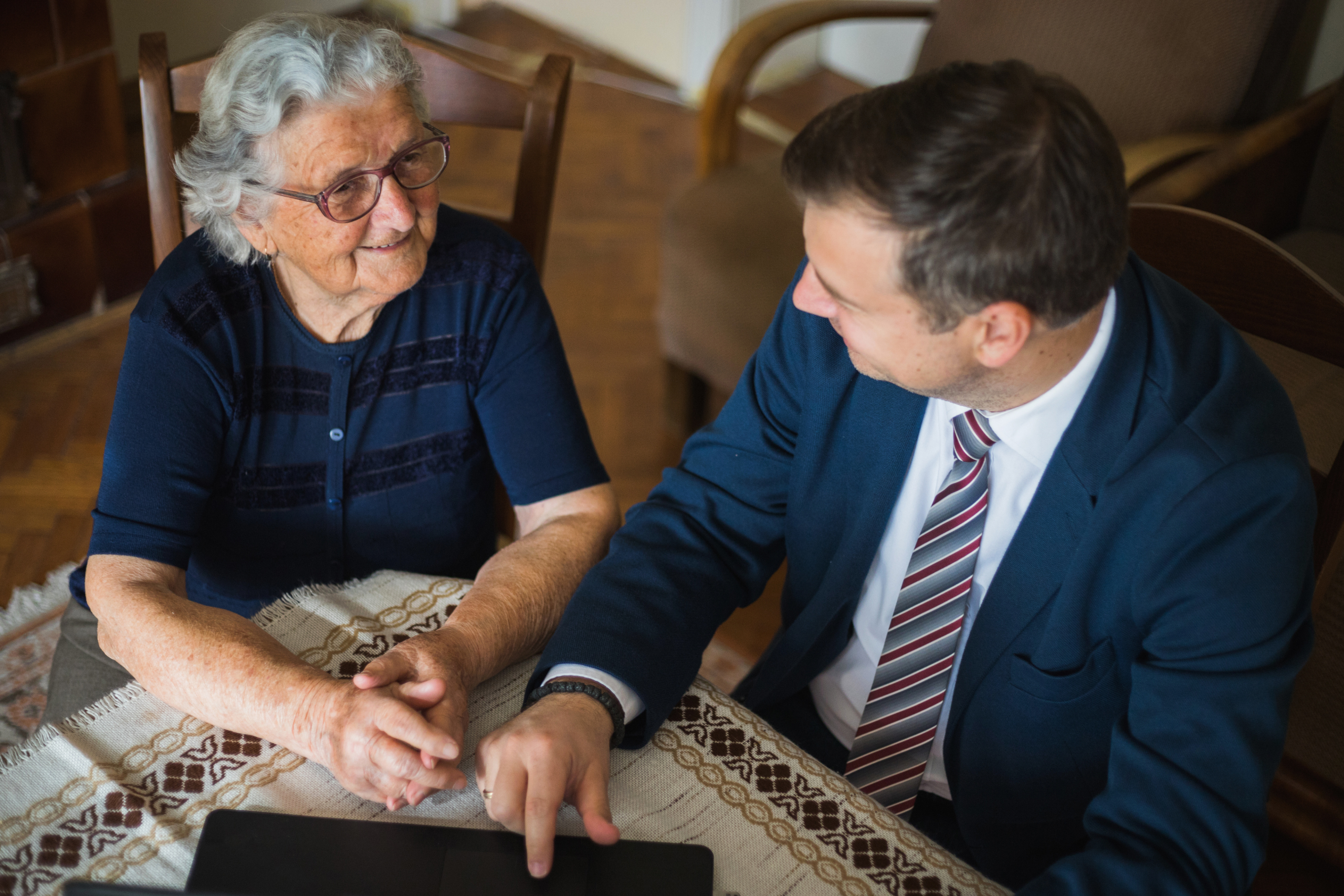 Elderly woman smiling and holding hands with a man in a suit, as he points to a document on a tablet during a legal consultation.