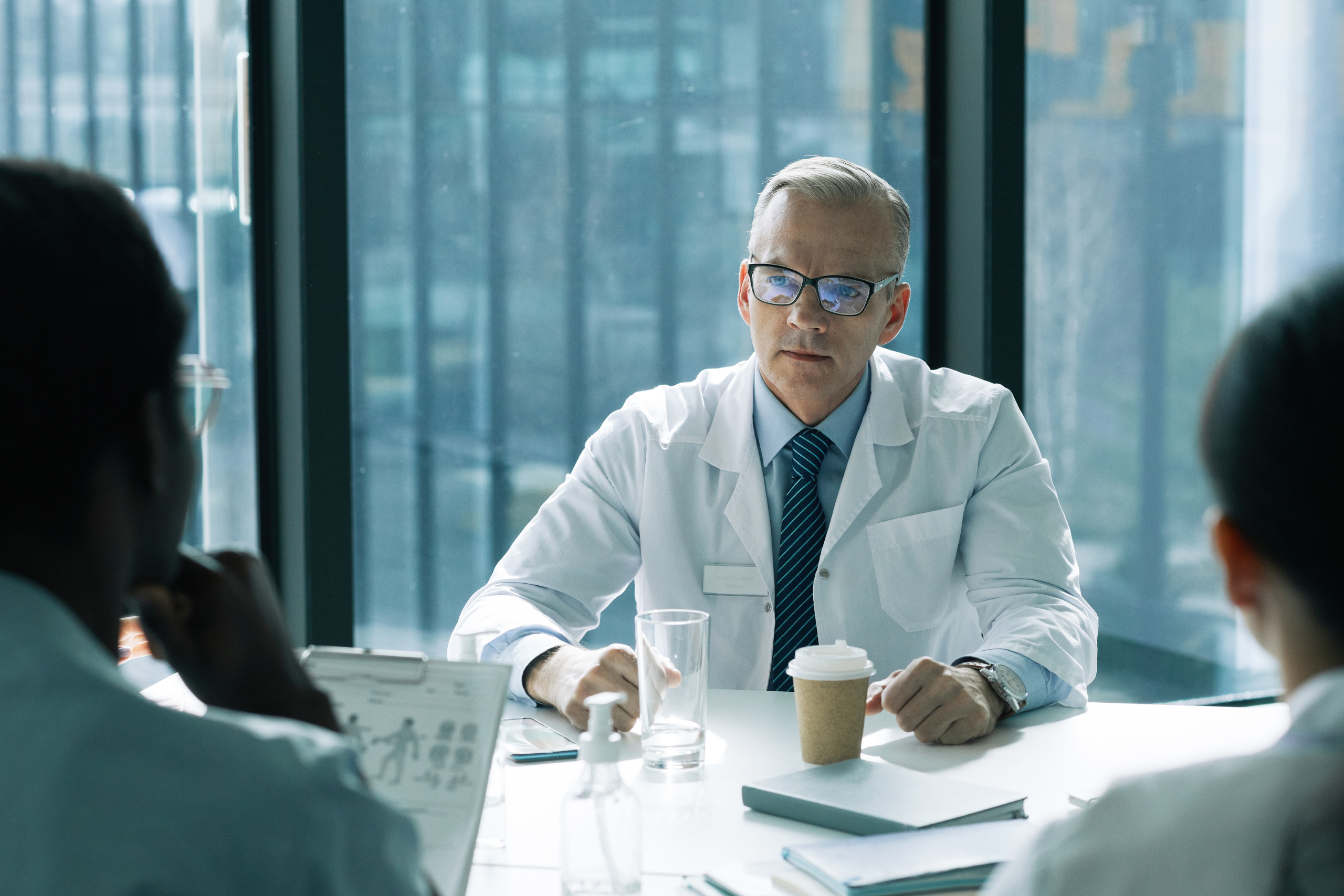 Middle-aged male doctor in a lab coat and glasses sits at a table with colleagues, discussing documents in a bright office setting.