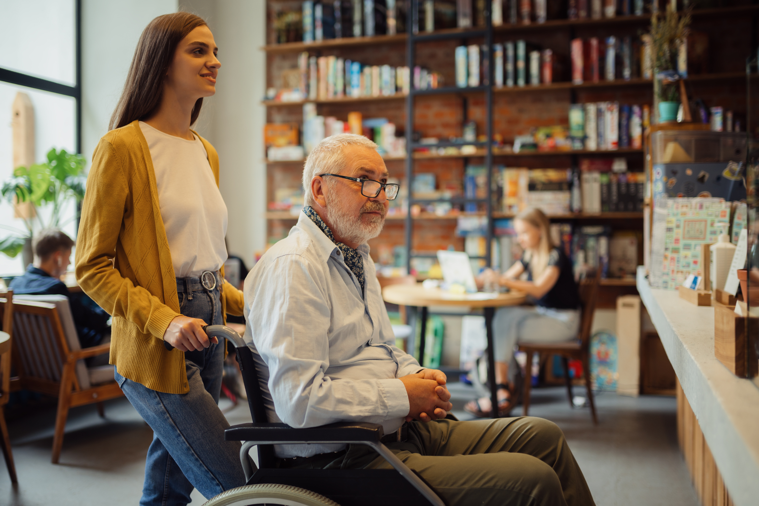 Young woman assisting an elderly man in a wheelchair through a cozy café, with shelves of books and patrons in the background.