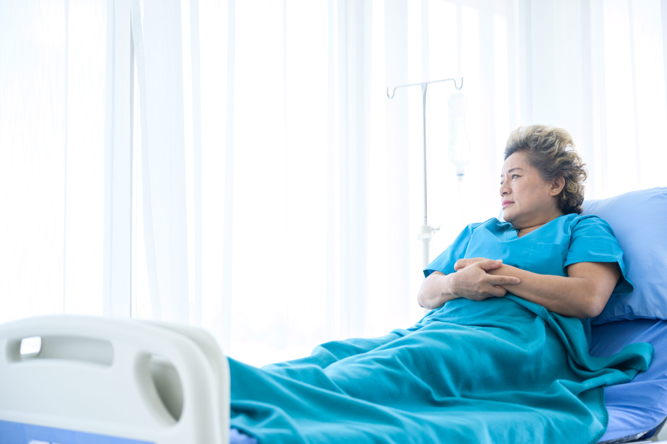 Elderly woman lying in a hospital bed, wearing a blue gown and gazing out the window, symbolizing nursing home neglect and isolation.