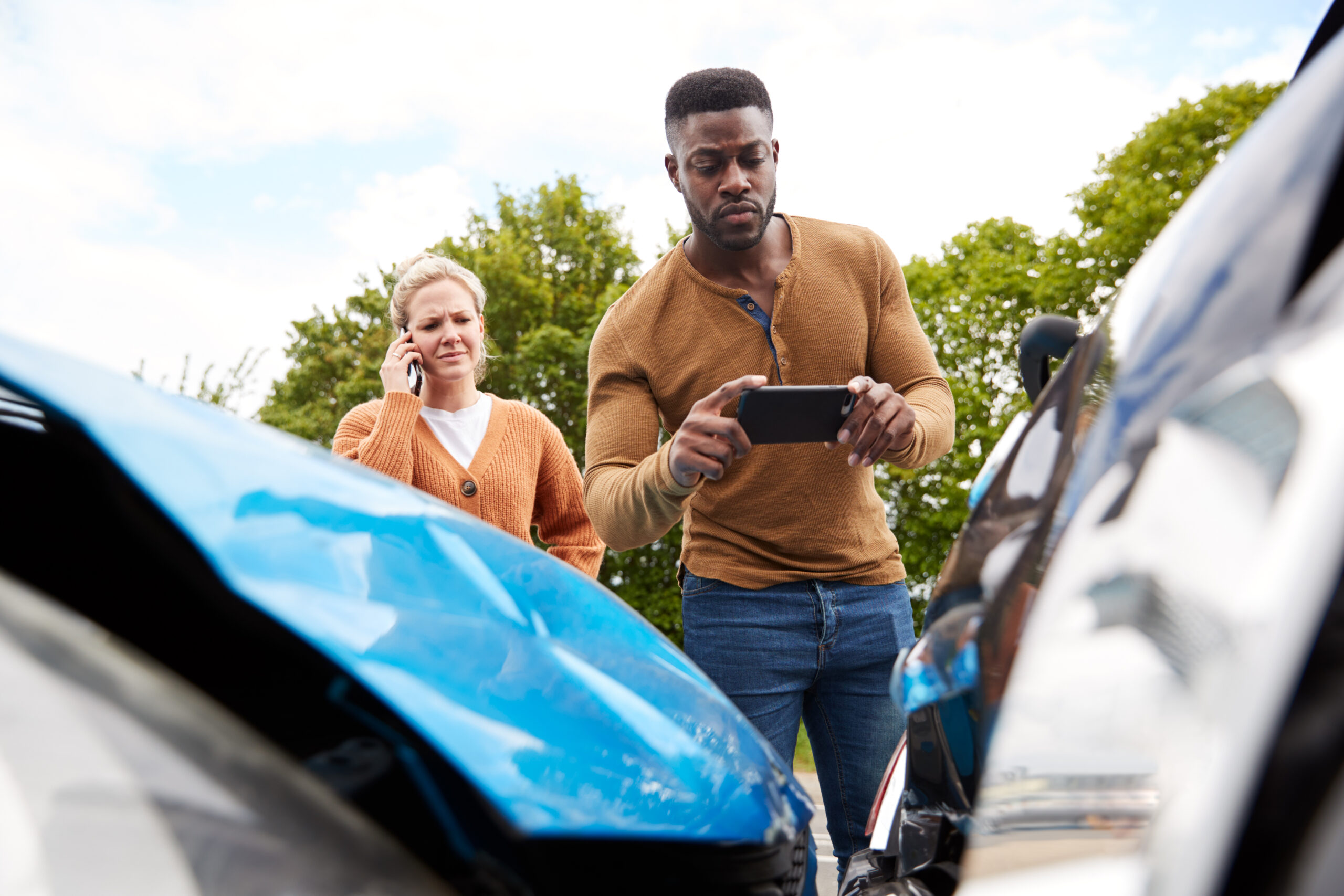 Man taking photos of car damage with his phone while a woman makes a phone call, emphasizing documentation after an accident.