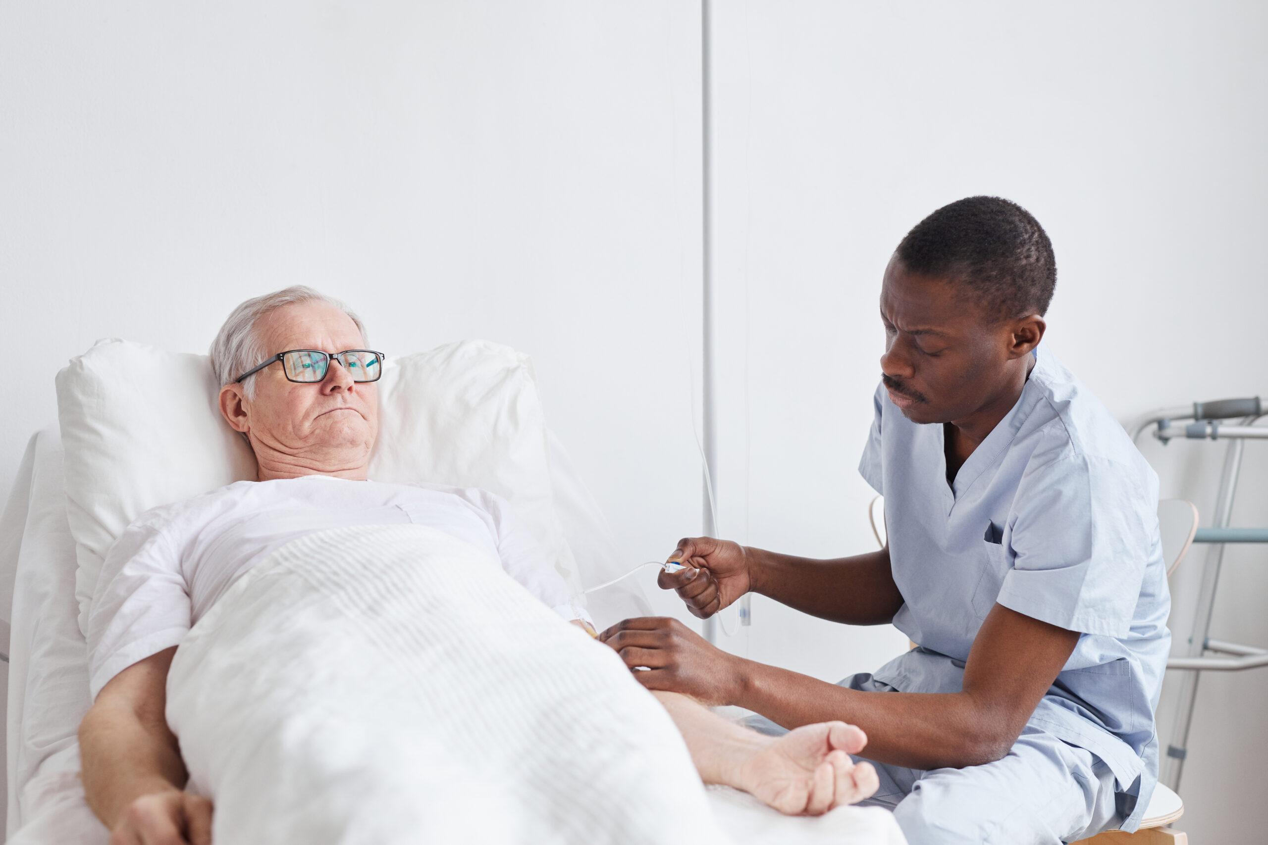 Elderly man lying in a hospital bed receiving care from a nurse adjusting his IV, illustrating the importance of attentive medical care.