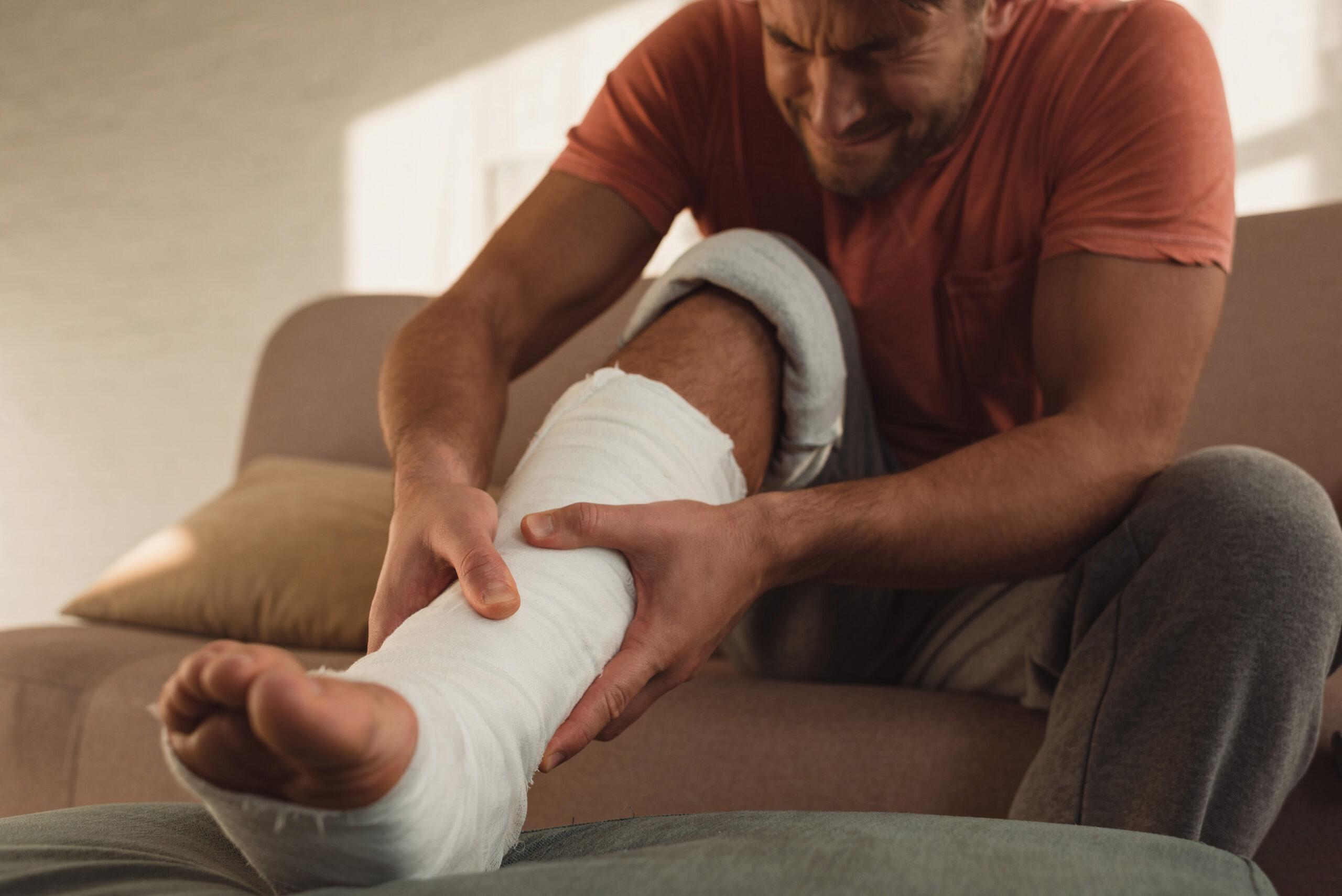 Man sitting on a couch holding his bandaged leg in pain, representing the challenges of recovering from an injury and navigating claims.