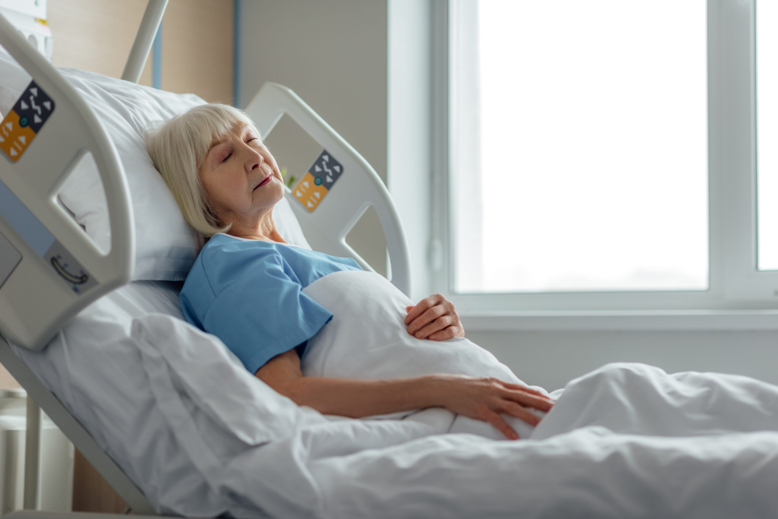 Elderly woman resting in a hospital bed near a bright window, highlighting vulnerability and the need for proper nursing home care.