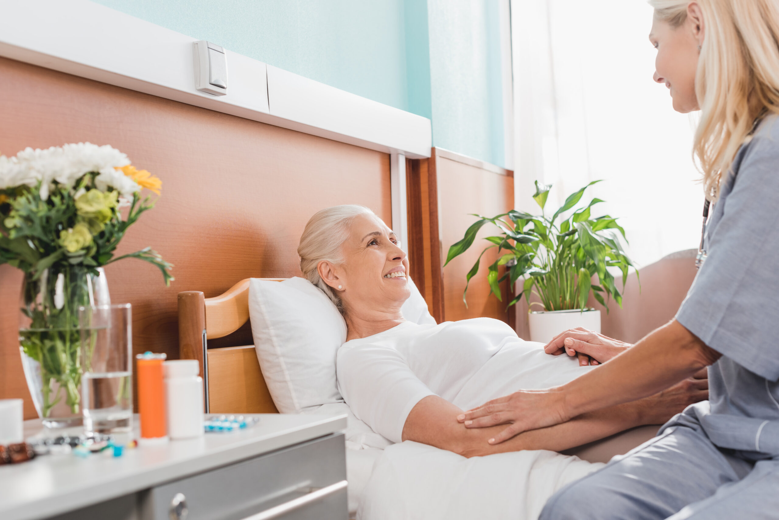 Smiling elderly woman in a nursing home bed receiving supportive care from a nurse, with flowers and medication on the bedside table.