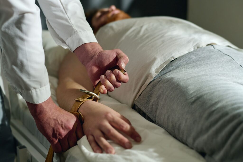Close-up of doctor’s hands fastening brown leather wrist restraint on nursing home resident’s arm in bed  
