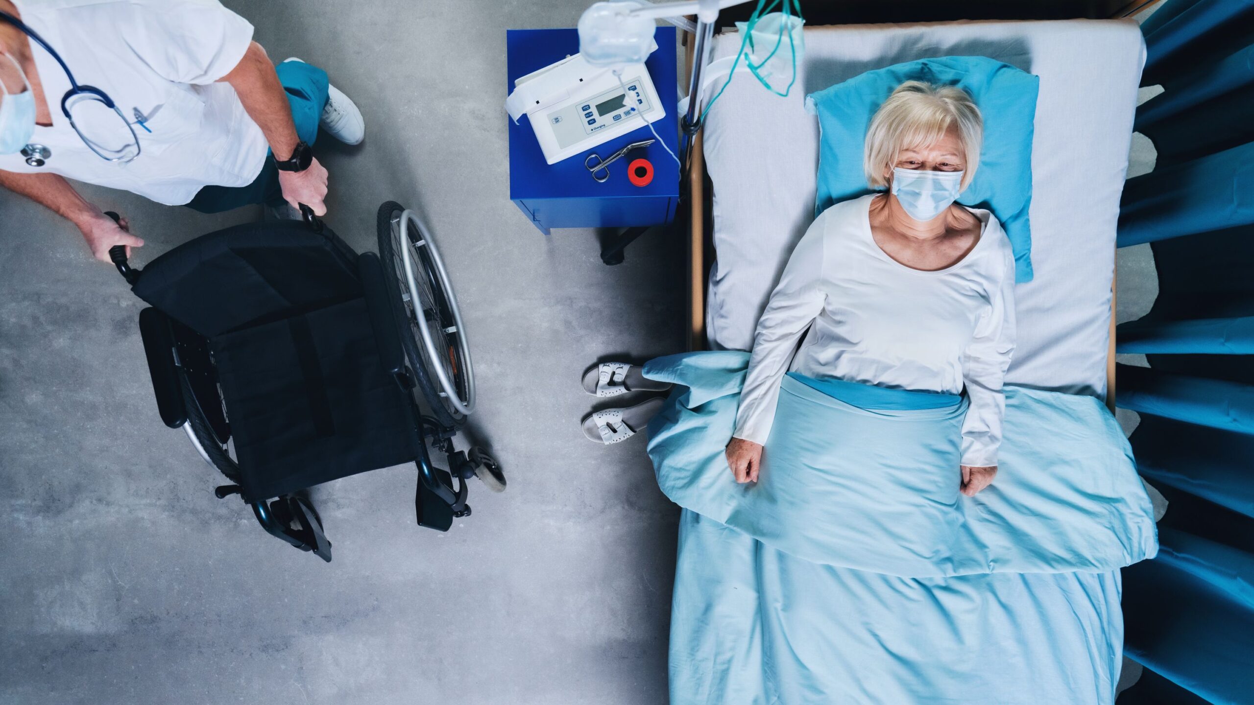 Overhead view of masked doctor arriving with wheelchair next to elderly nursing home resident lying in bed