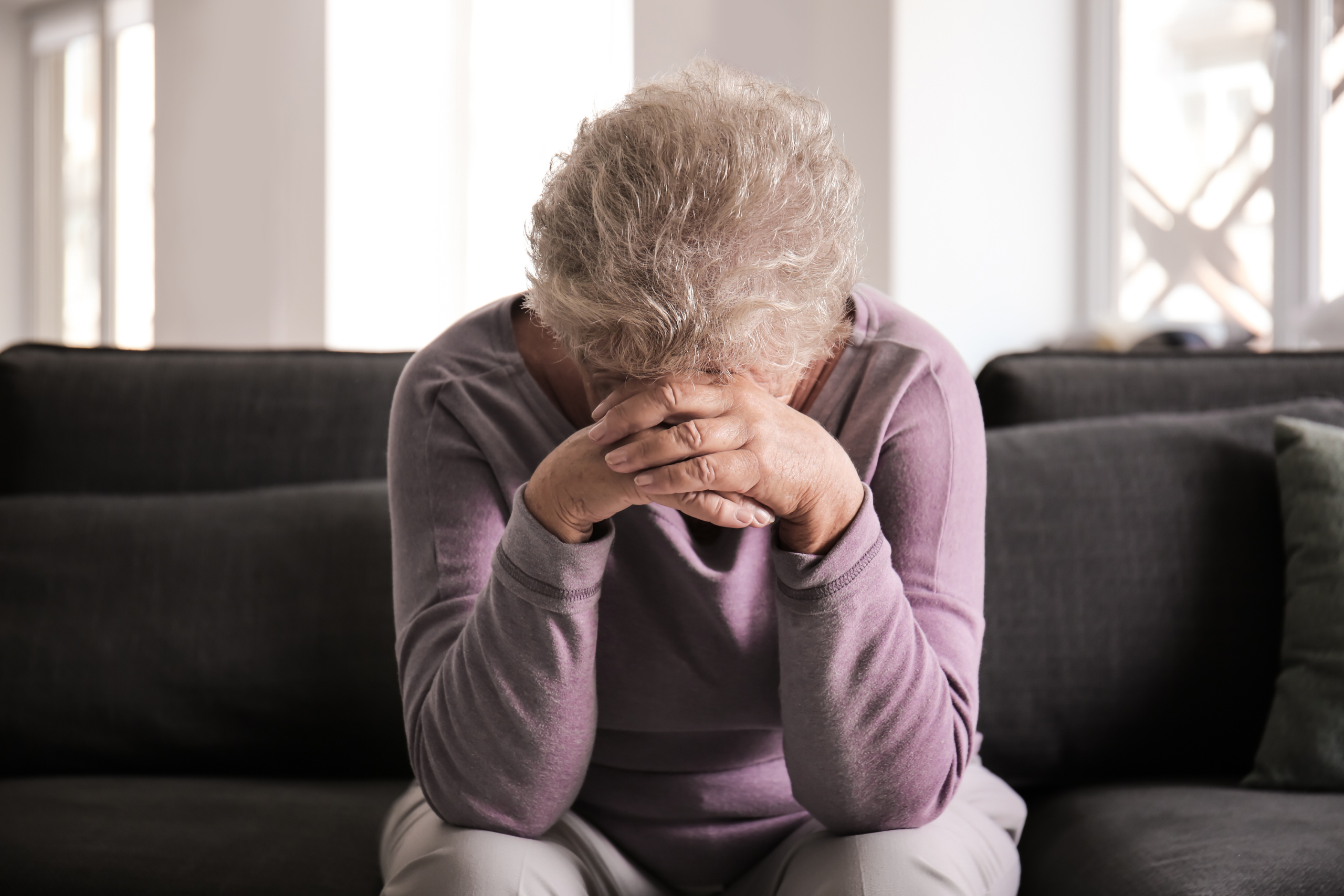 Distressed elderly woman sitting with head in hands on a couch.