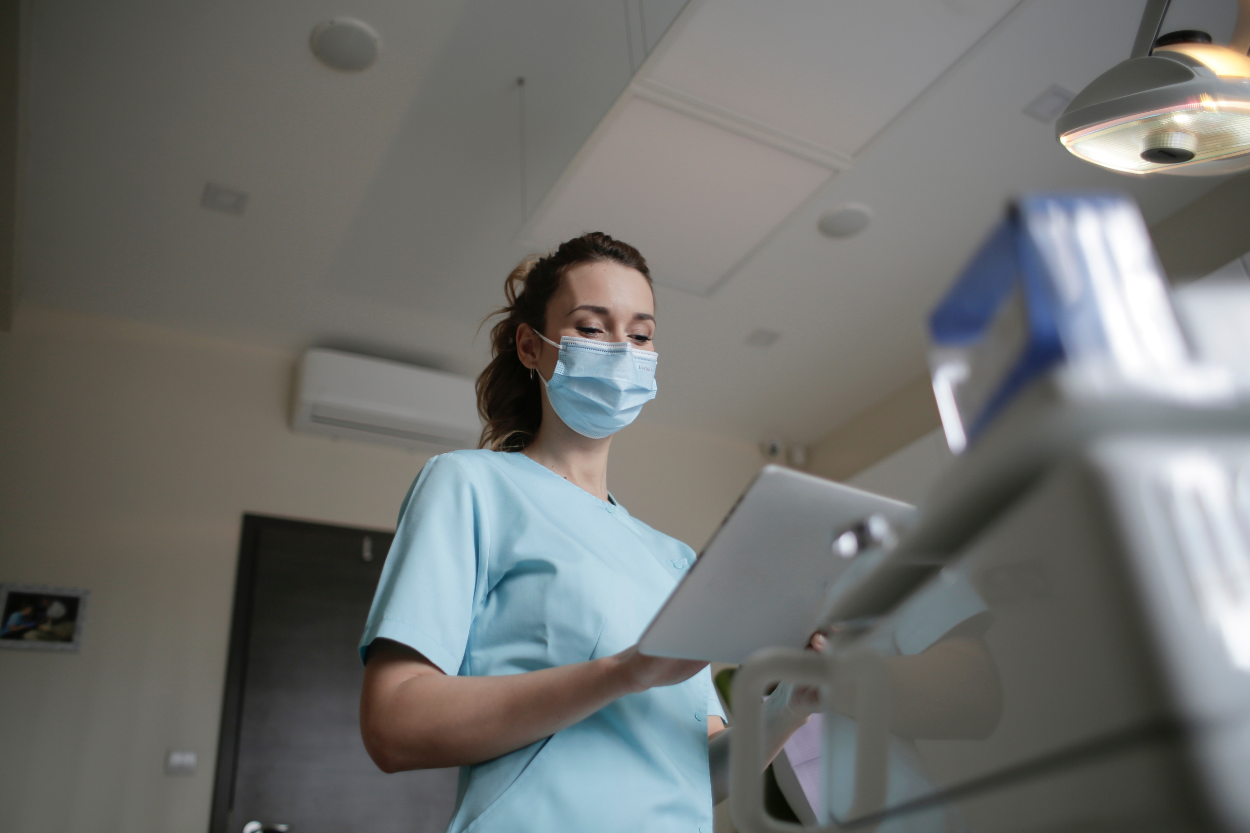 Nurse in scrubs and mask reviews a chart on a tablet in a medical room.