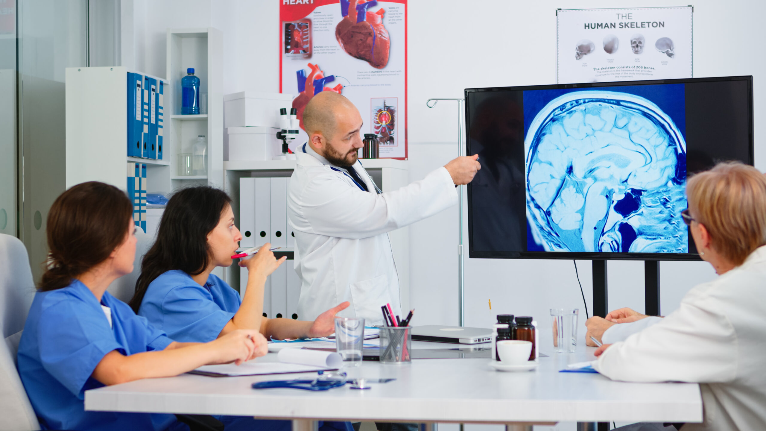 A doctor presenting a brain scan on a screen to a group of medical professionals during a discussion in a hospital meeting room. 