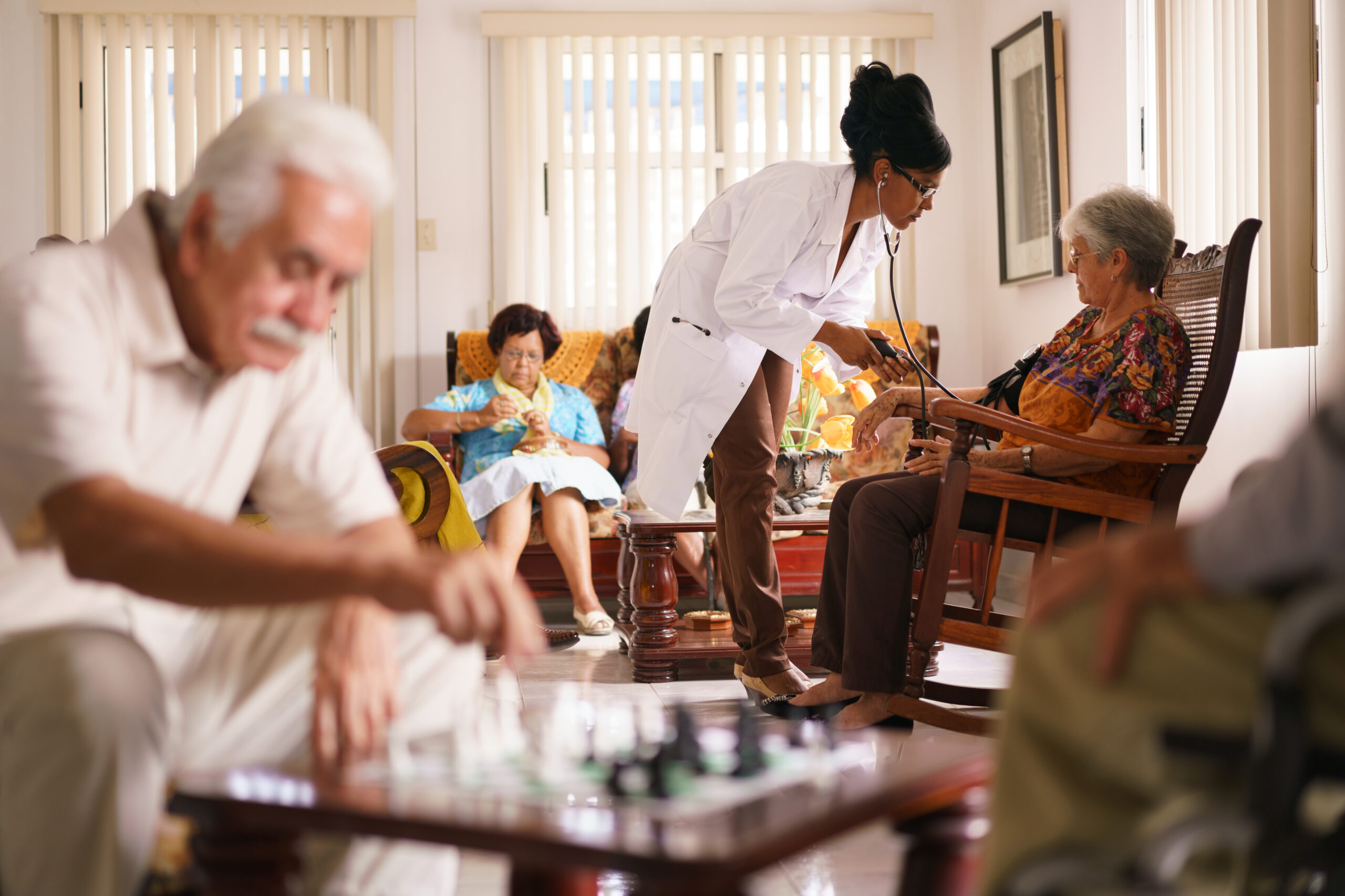 A doctor checks an elderly woman's blood pressure in a nursing home while other residents engage in activities like chess and knitting. 