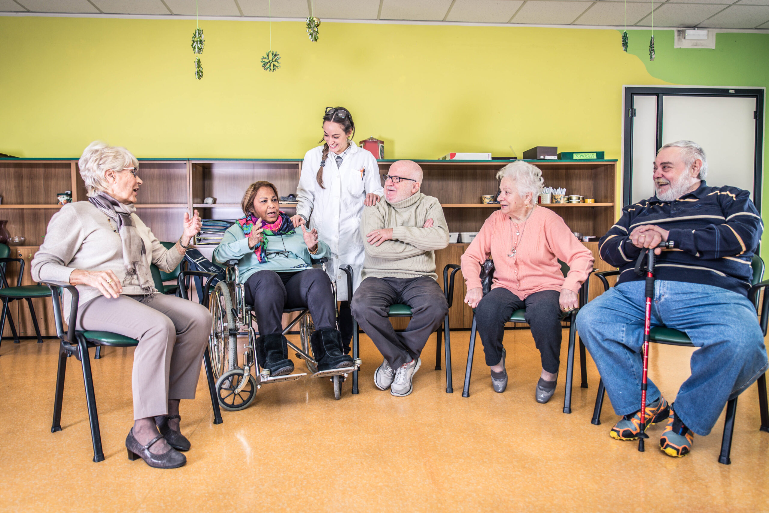 A group of elderly nursing home residents, including a woman in a wheelchair, engage in a cheerful conversation with a caregiver in a bright common room. 