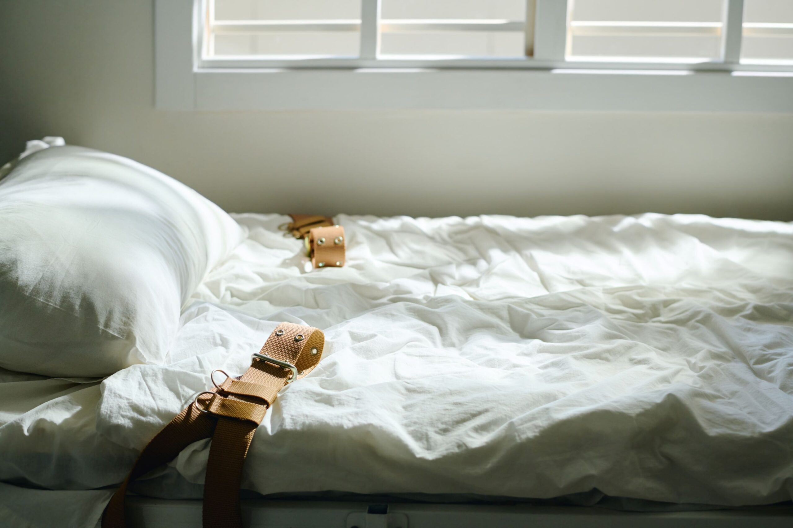Side view of white bed with brown leather restraints secured to mattress under window blinds in a bright room 