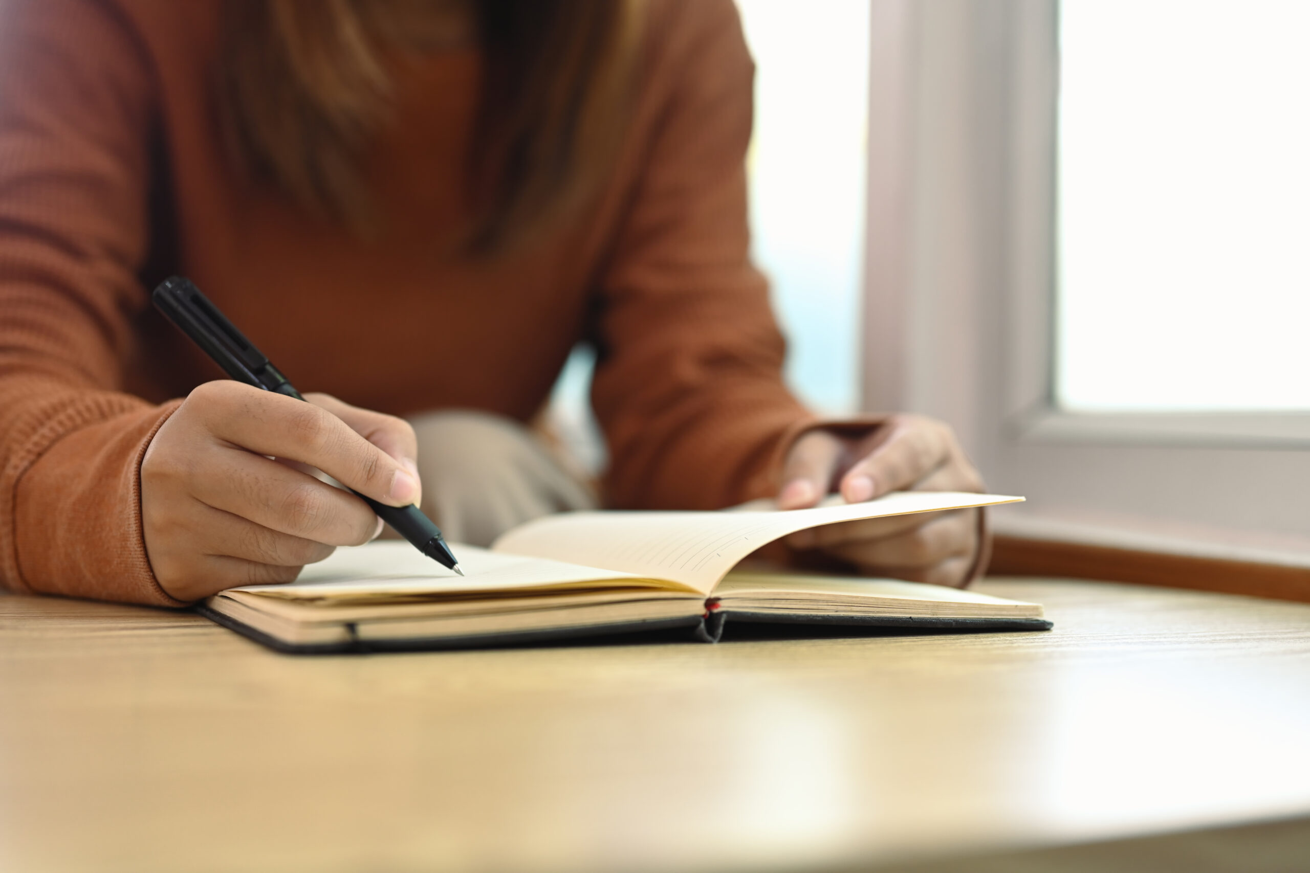 A person in a brown sweater writing in a pain journal on a wooden table, documenting symptoms, triggers, or pain management notes. 