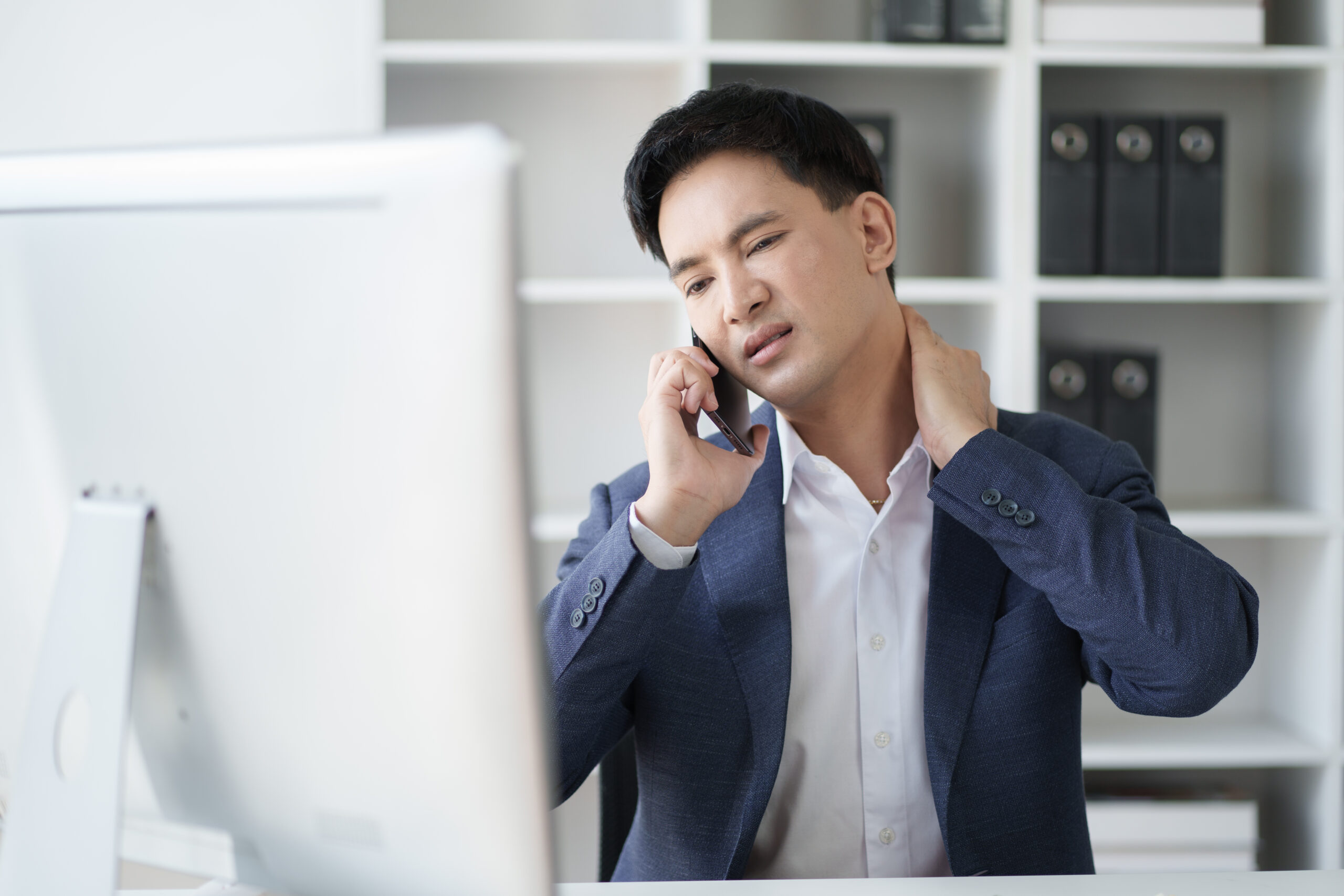 A man in a suit holding his neck in pain while talking on the phone at his desk, suggesting discomfort or possible injury.