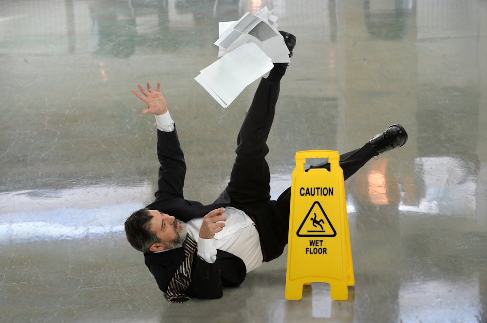 A man in formal attire is falling backwards on a shiny floor, scattering papers. A yellow caution sign indicating "WET FLOOR" stands nearby in a bright environment.