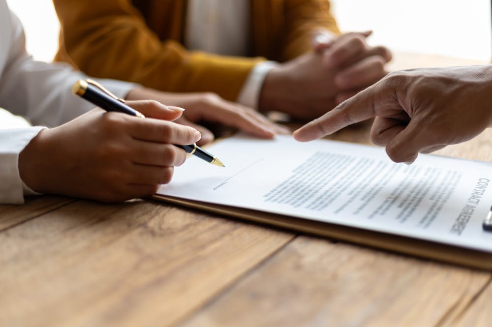 A hand holding a pen writes on a document, while another hand points at the text. The setting is a wooden table, suggesting a formal discussion or agreement.