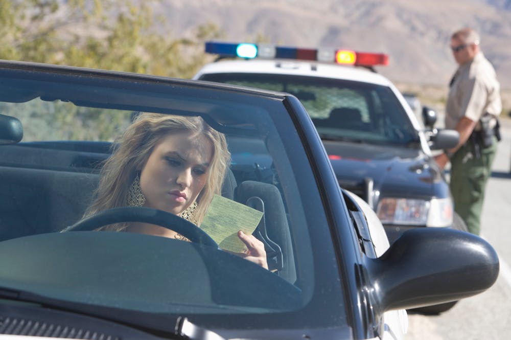 A woman in a car looks at a traffic ticket while a police officer approaches from behind; police car lights are on, indicating a traffic stop in a desert-like area.