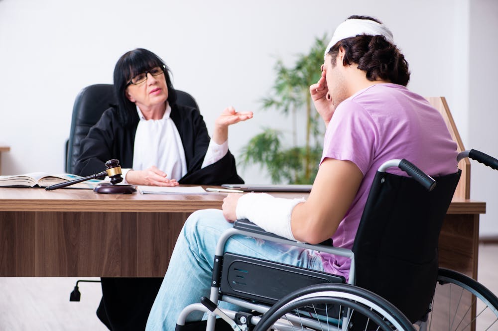 A judge, in a black robe and glasses, gestures while seated at a desk. A man in a wheelchair, with bandages on his head and arms, listens intently in a well-lit office.
