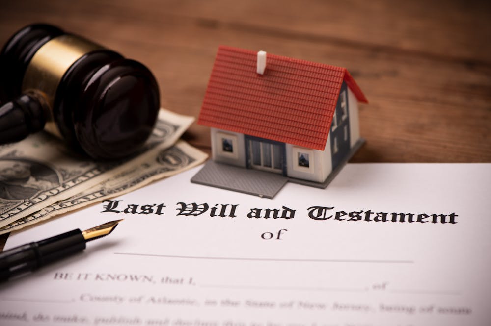 A miniature house sits on a document labeled "Last Will and Testament," accompanied by a gavel and a fountain pen, with dollar bills in the background, suggesting a legal context of inheritance.