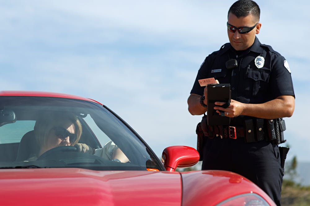 A uniformed police officer examines a device while standing by a red car, with a driver inside, against a clear sky backdrop.